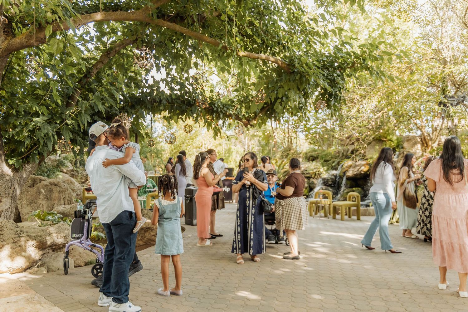 Guests mingle outdoors under lush green trees at a casual daytime gathering on a sunny patio.
