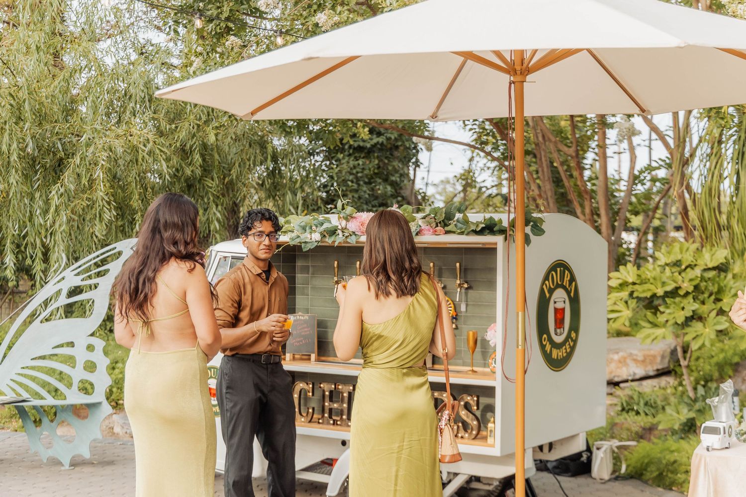 Two women in yellow dresses ordering drinks at an outdoor mobile bar cart with umbrella shade.