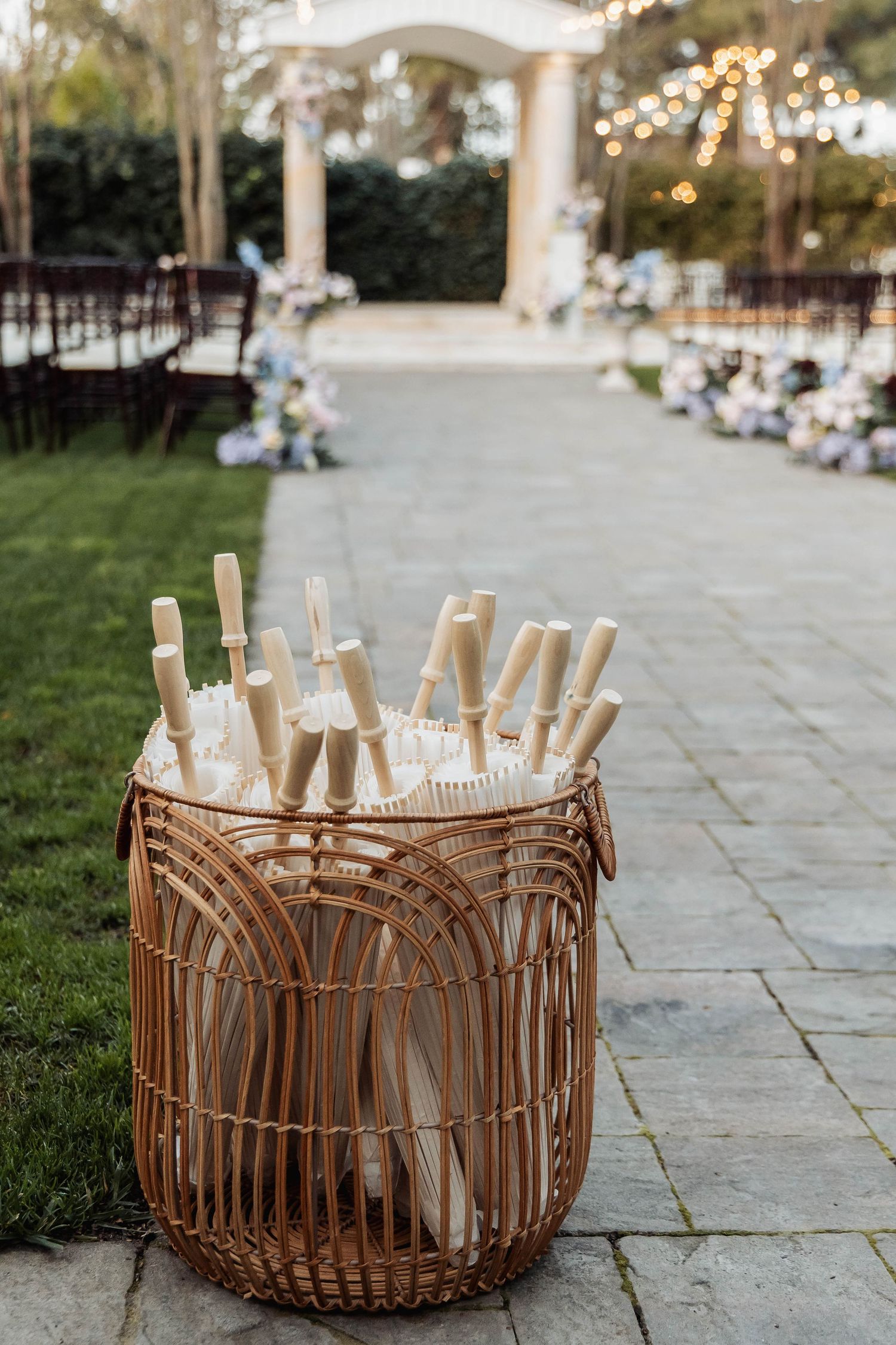 Wicker basket filled with handheld fans at an outdoor wedding ceremony with string lights and floral aisle decorations.