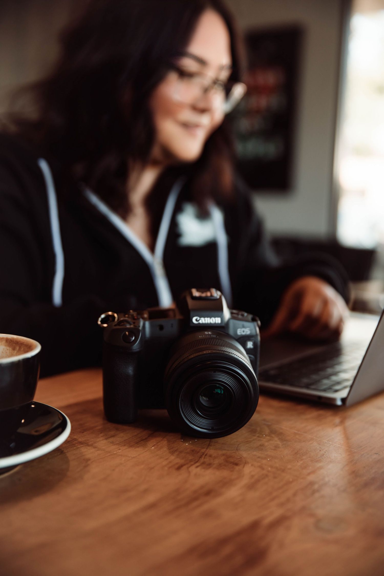 Woman photographer working on laptop beside Canon EOS camera and coffee cup on wooden table.