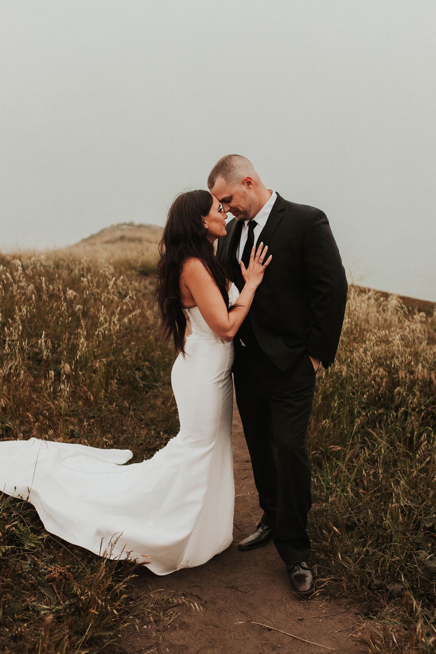 A wedding couple poses together on a golden hillside trail during sunset.