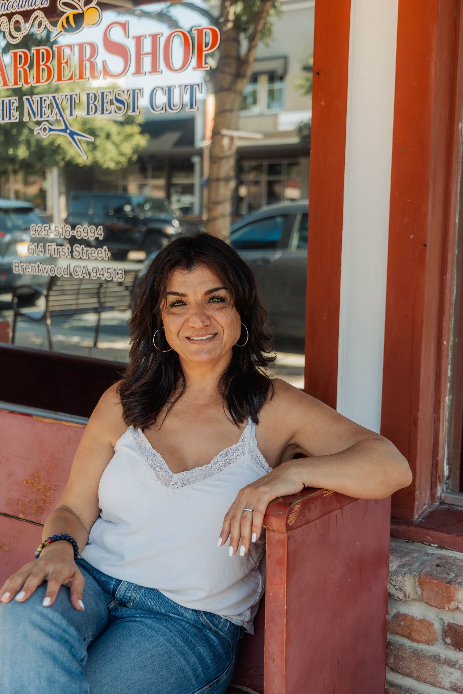 Casual portrait of someone in white top and jeans sitting on red bench outside barber shop.