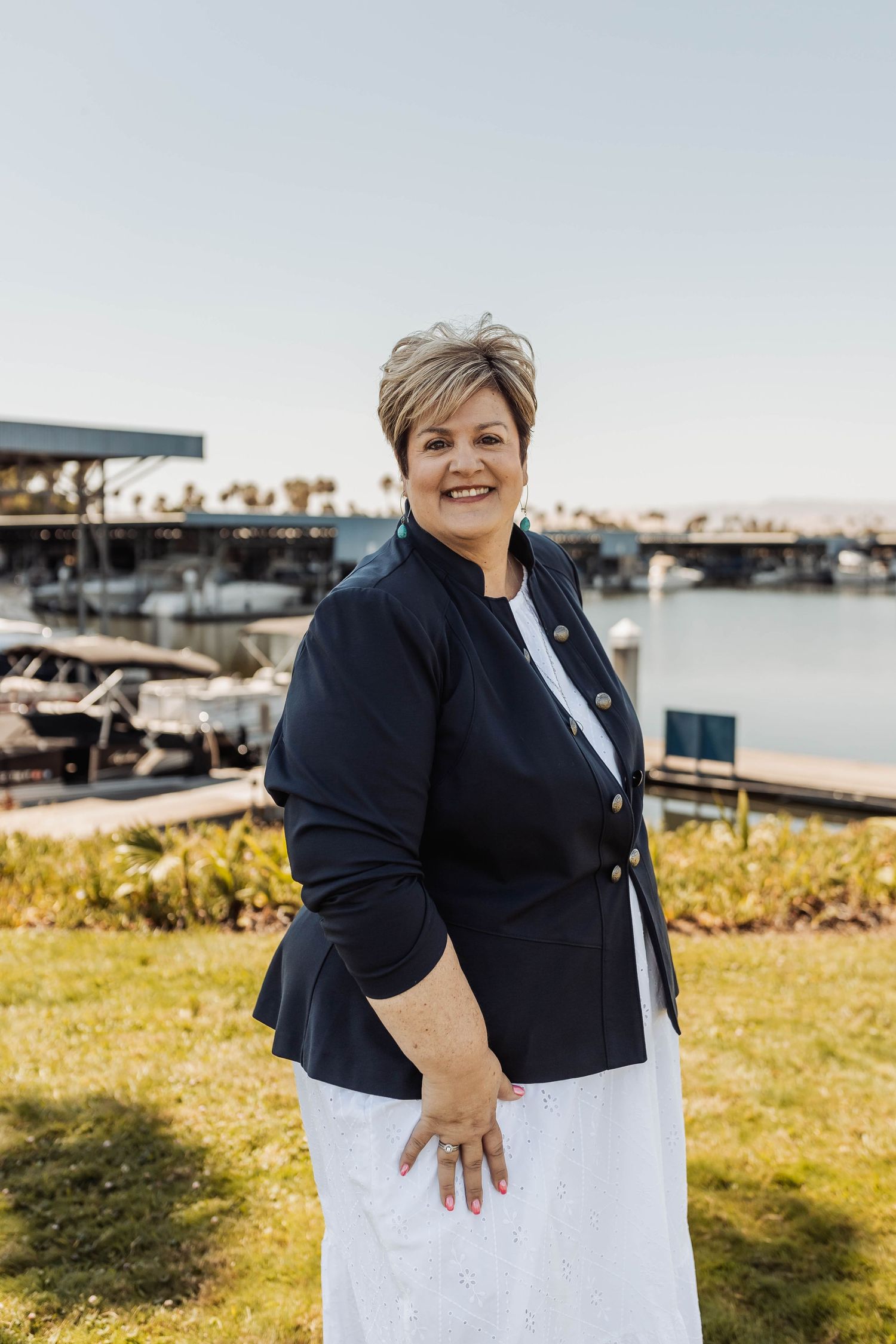 Professional in navy blazer and white dress poses outdoors near a marina on a sunny day.