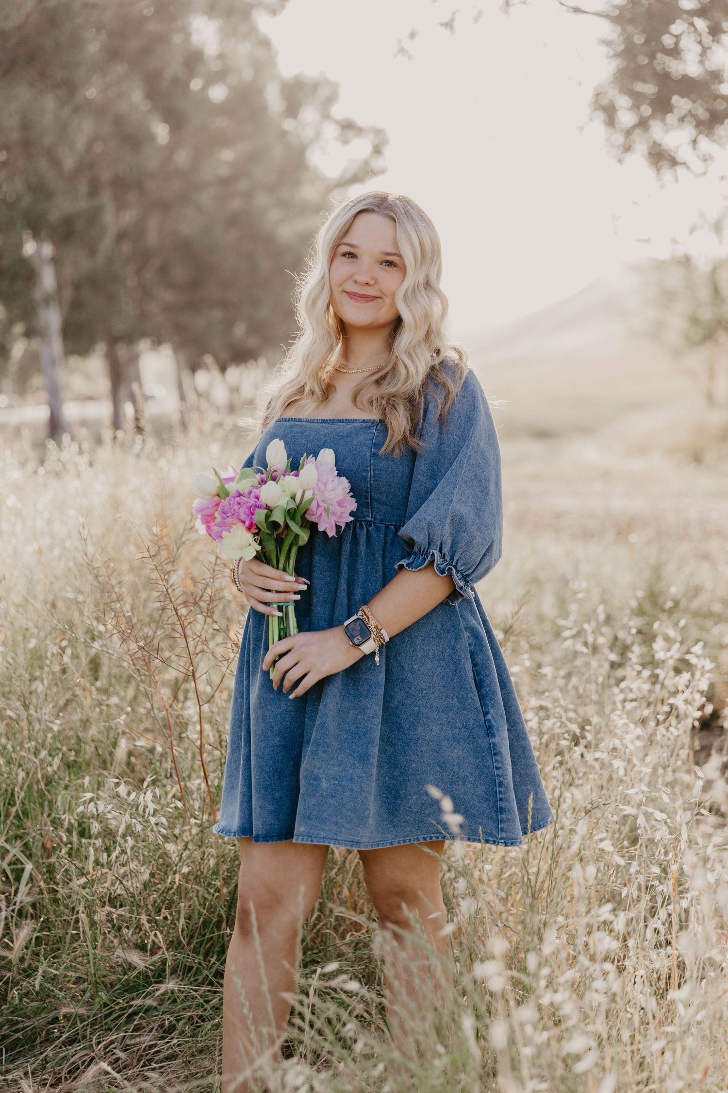 Woman in denim dress holding pink flowers in a sunlit meadow of tall grass.