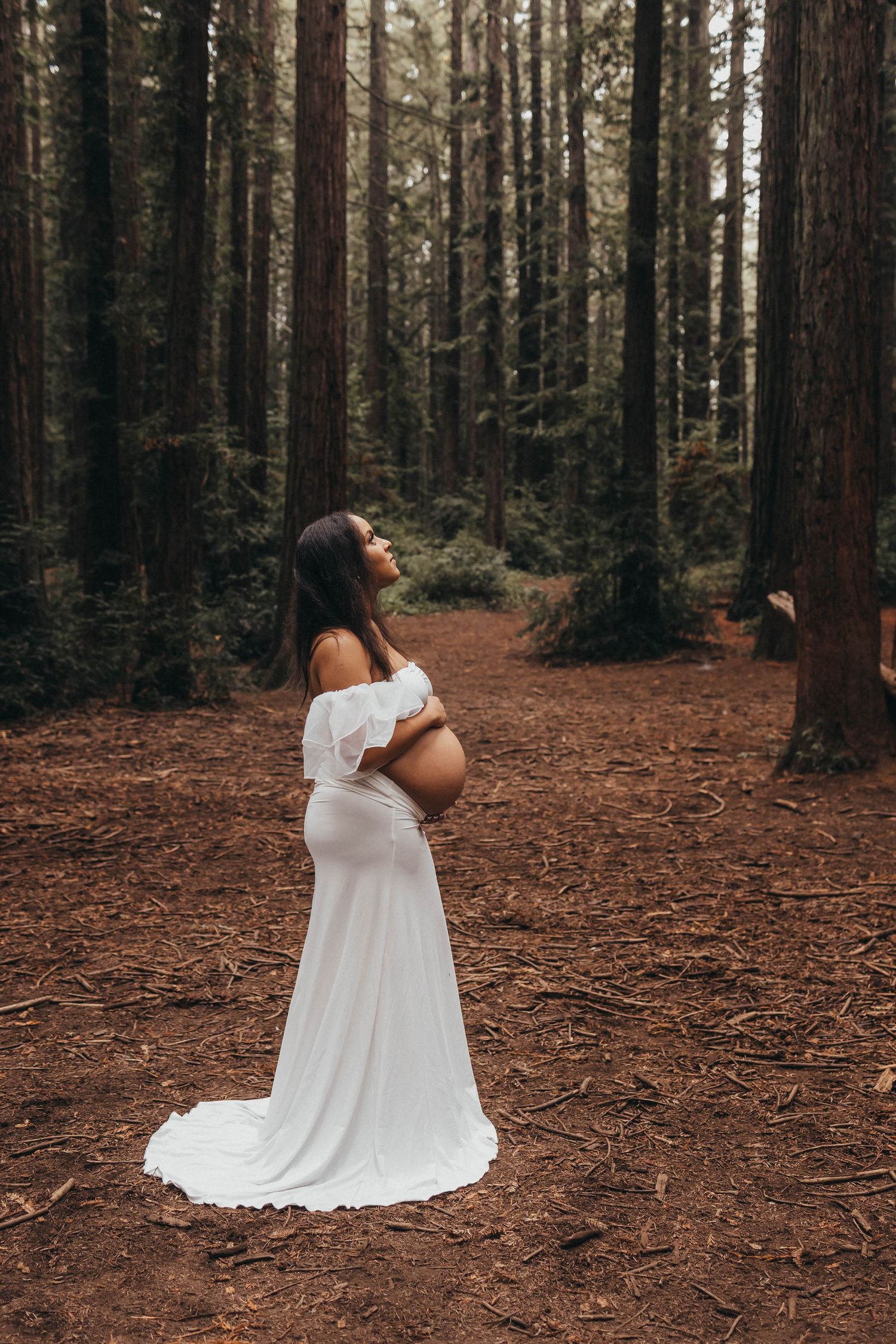 Person in flowing white dress poses dramatically in a moody forest setting.