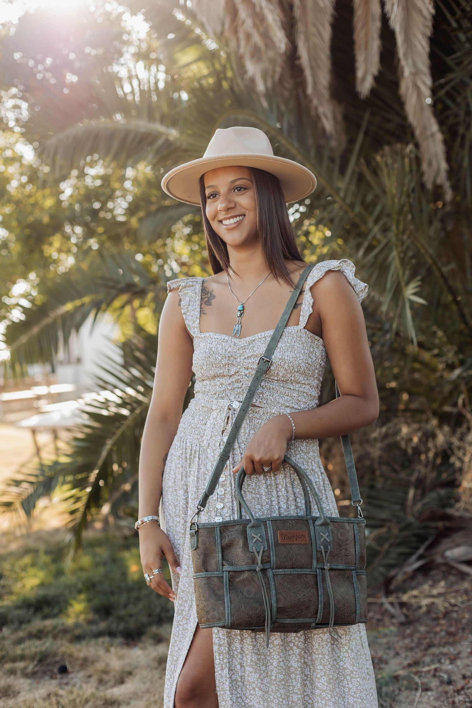 Woman in floral dress and sun hat poses outdoors with vintage leather bag in natural lighting.