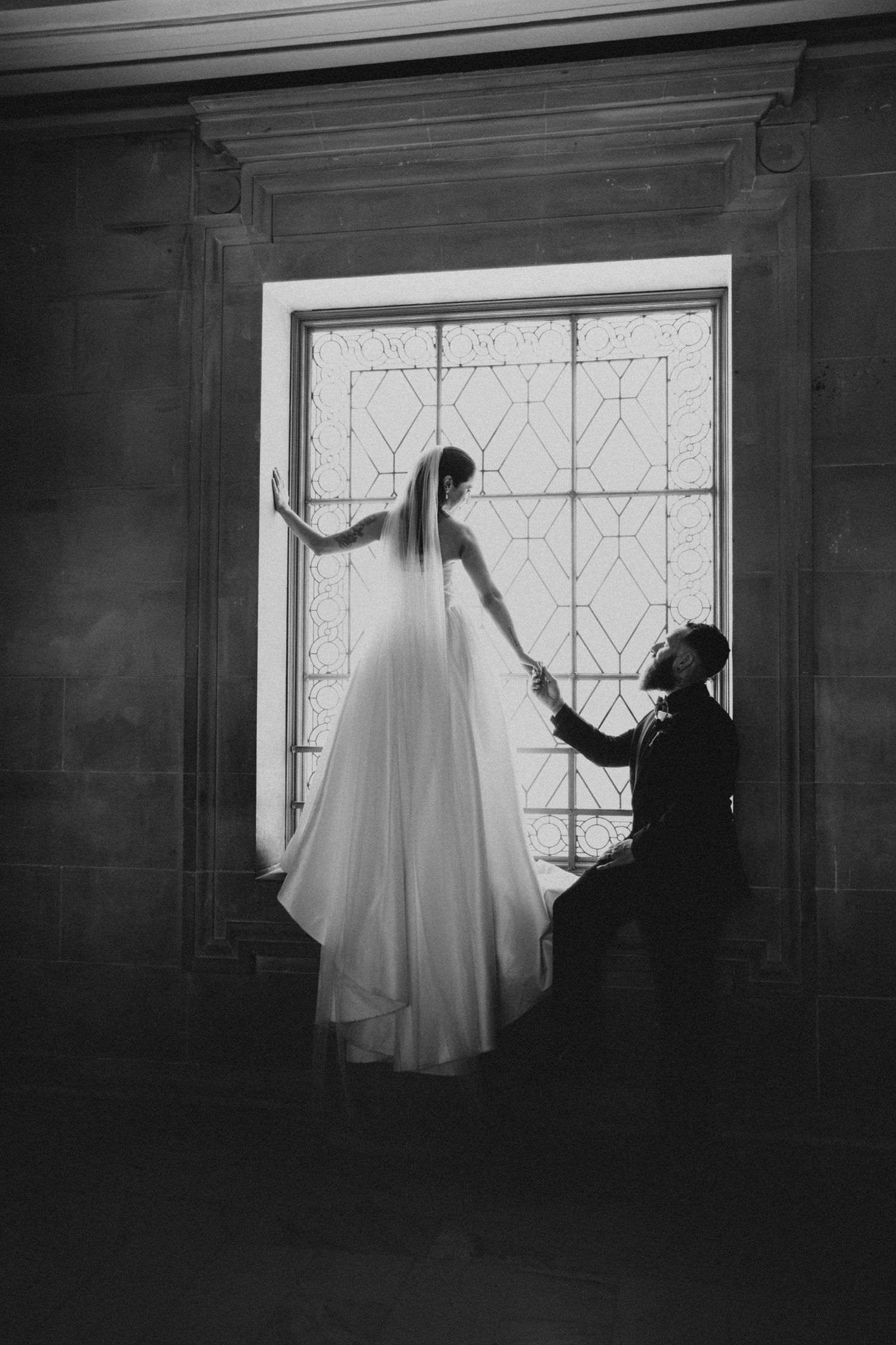 Black and white wedding photo of bride standing in window holding groom's hand as he kneels beside her.