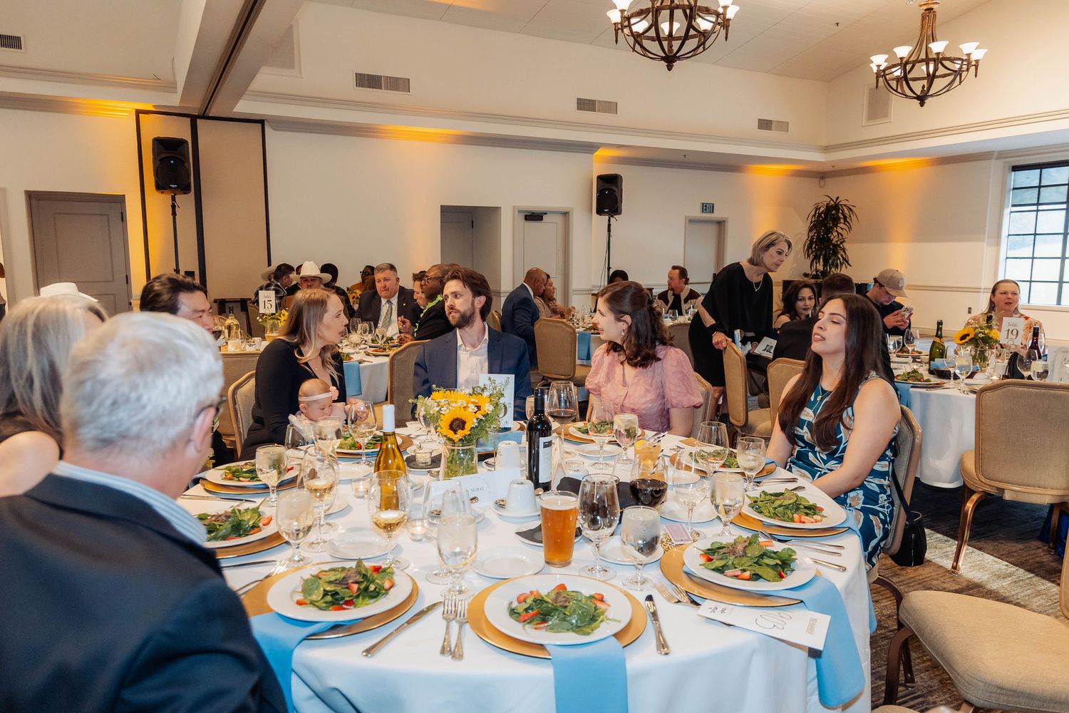 Elegantly dressed guests enjoy a formal dinner at round tables adorned with sunflowers and white linens.