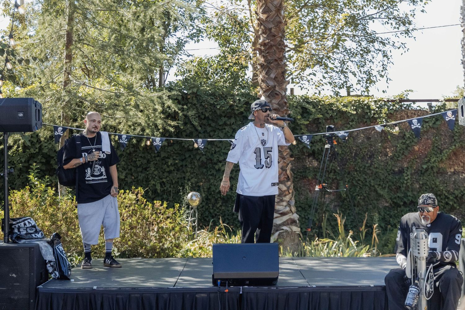 Three performers on an outdoor stage at a daytime event, surrounded by trees and string lights.
