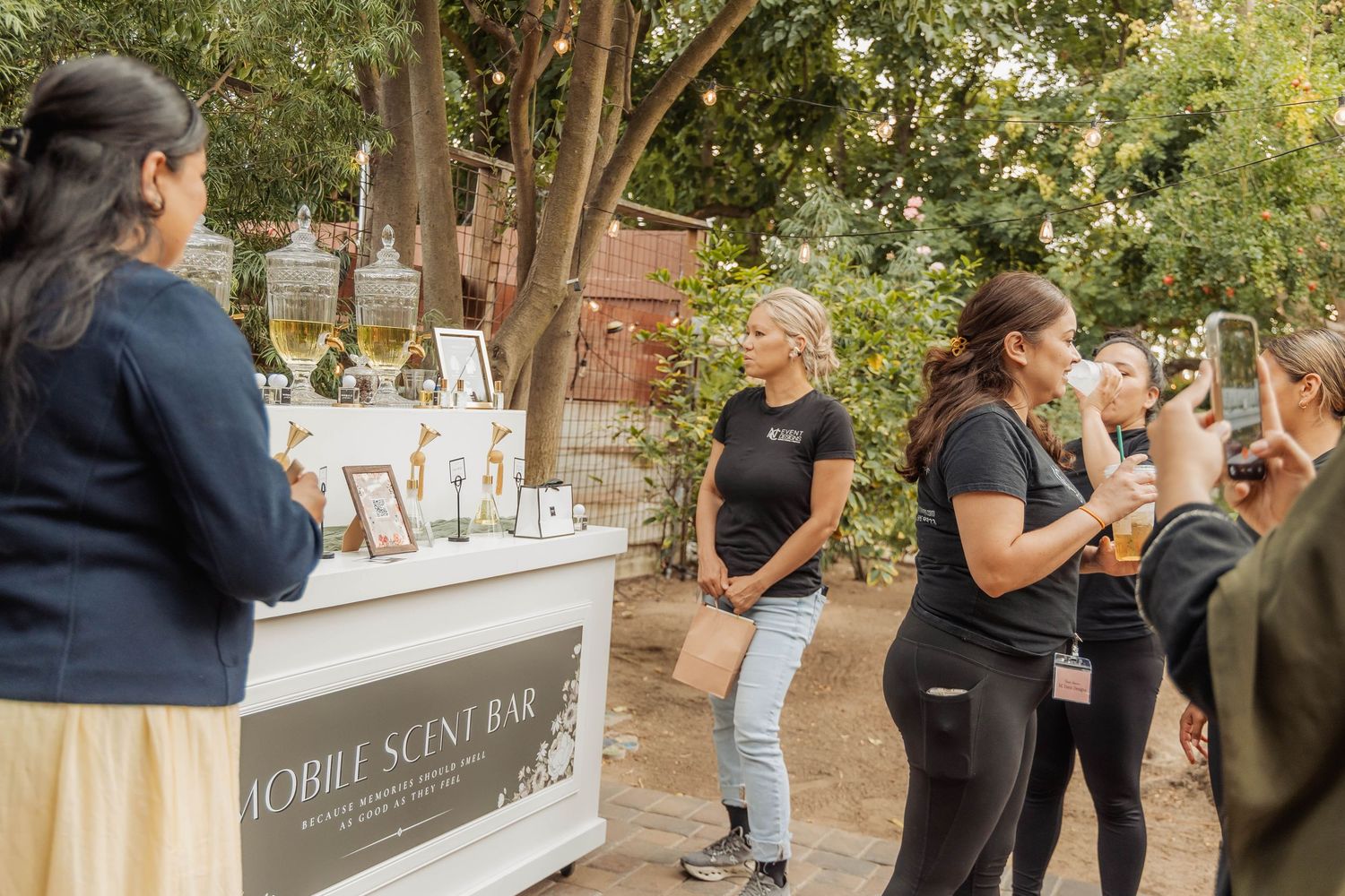 Women gather around a Mobile Scent Bar outdoor event, with one guest photographing the experience in a garden setting.