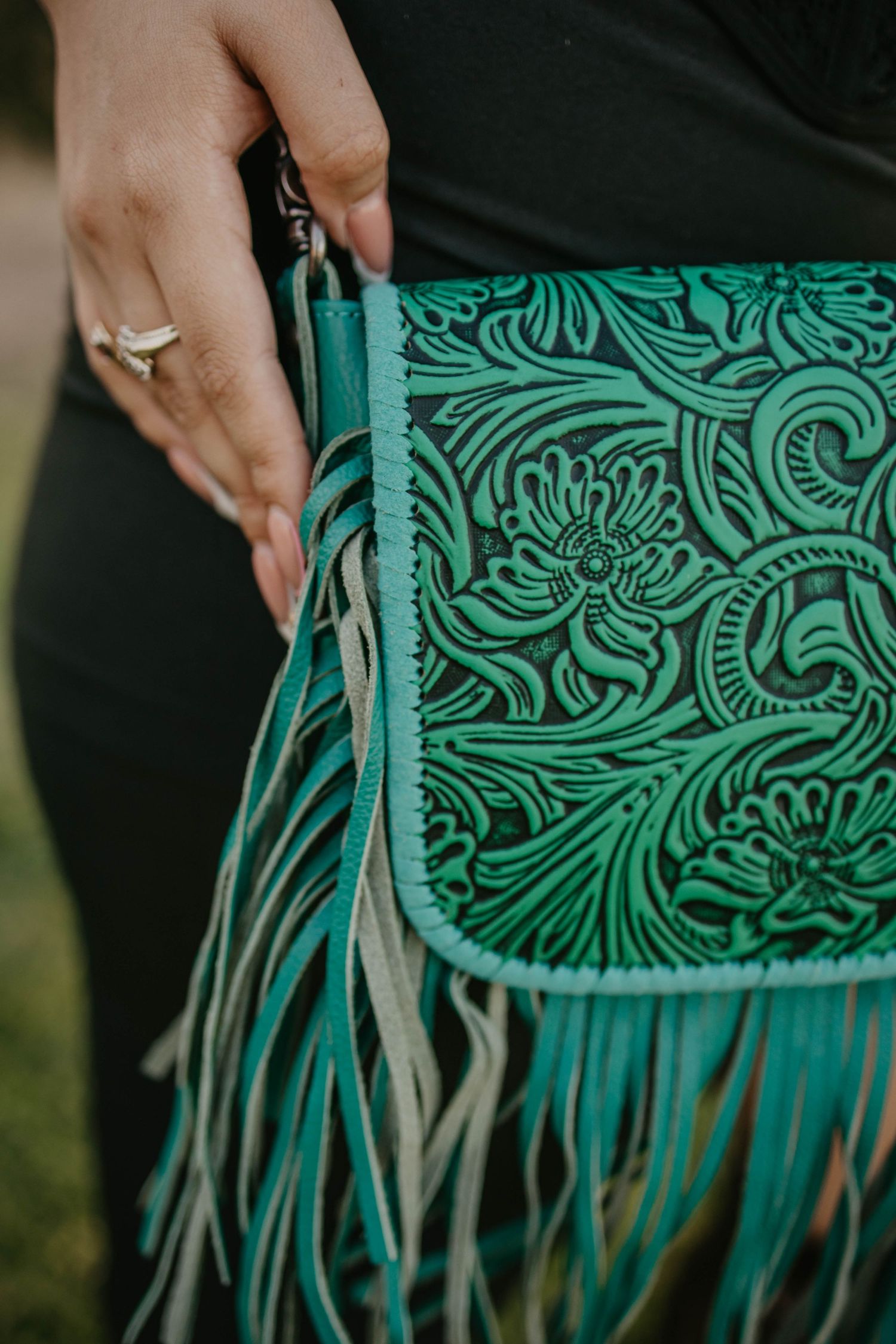 Close-up of a turquoise leather handbag with ornate floral tooling and fringe detail, with a hand holding the strap.