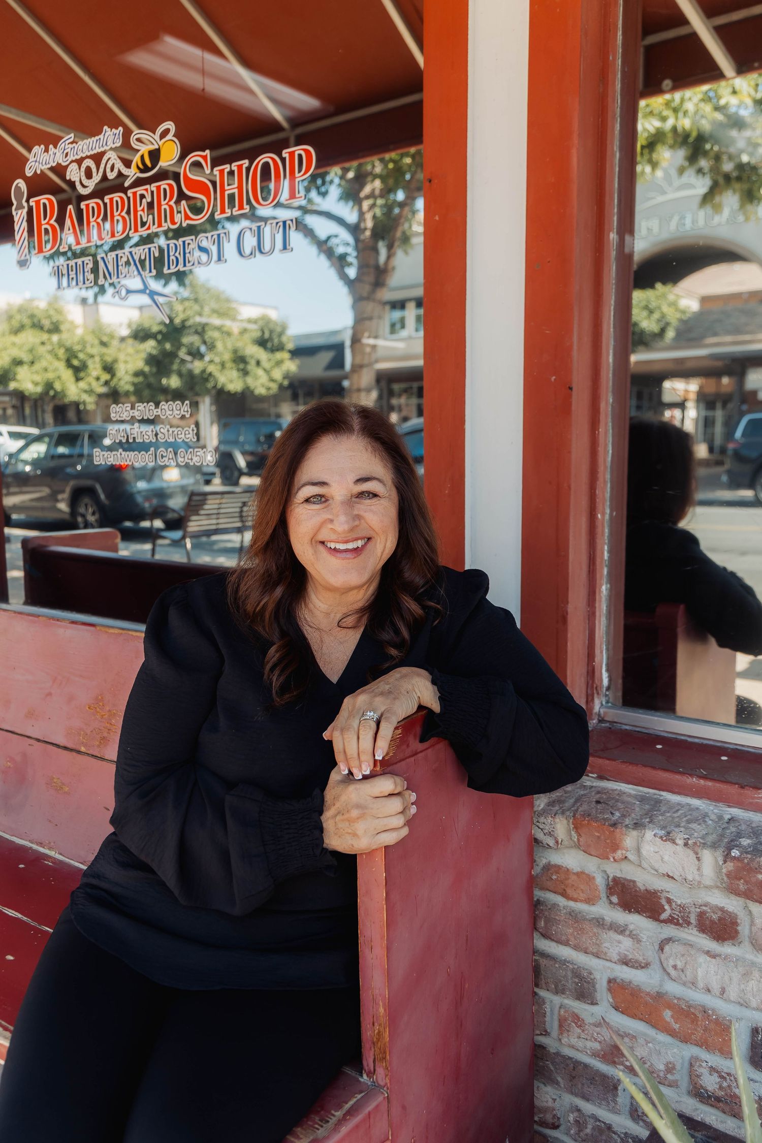 Smiling portrait of someone in black attire leaning against a red bench outside a barber shop.