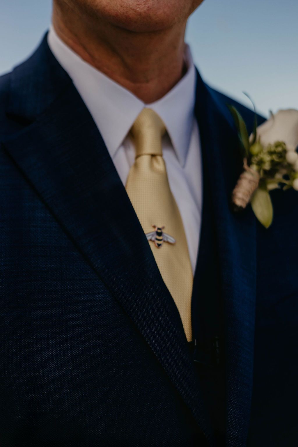 Close-up detail of navy suit with gold tie and boutonniere against blue sky.