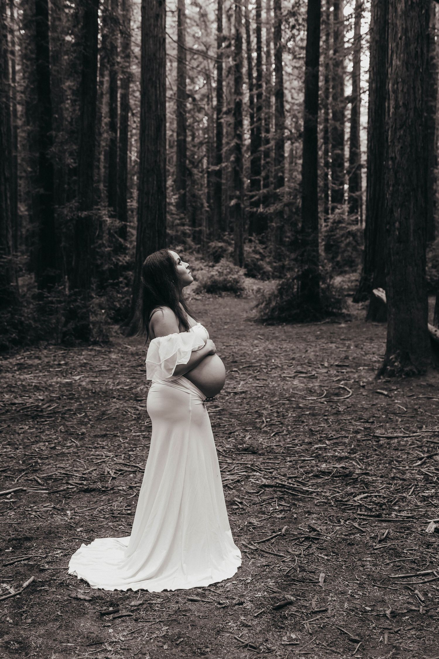 Pregnant woman in white gown standing in forest, black and white maternity photography among tall trees.