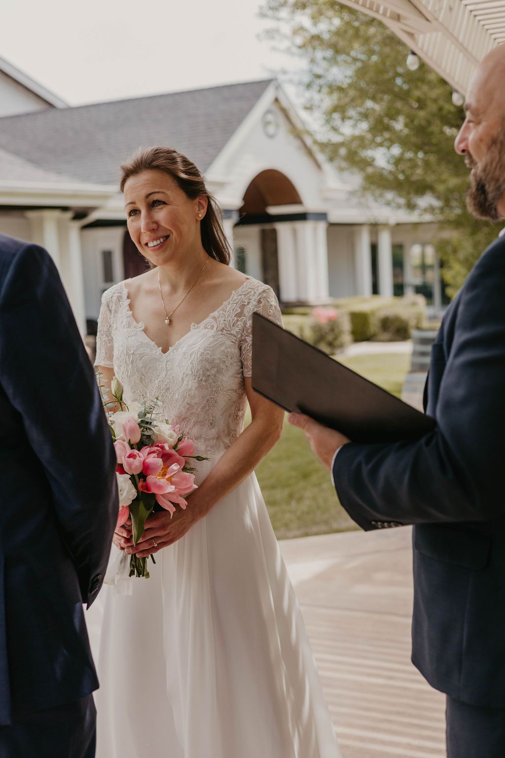 An intimate outdoor wedding ceremony with the bride holding a pink bouquet on a porch of a white building.