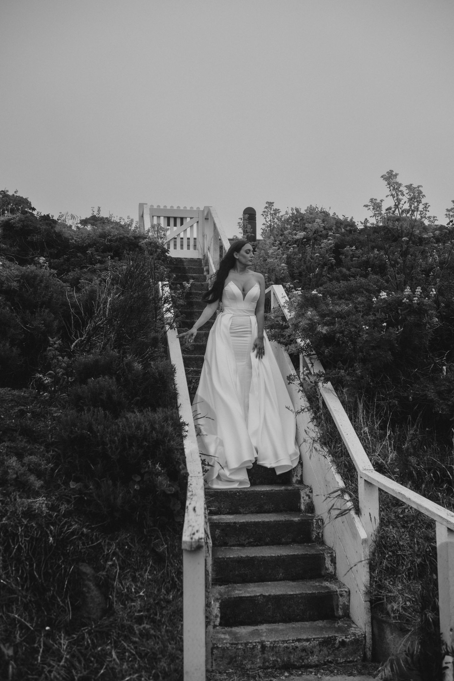 Black and white photo of a bride descending weathered white stairs in a flowing gown.