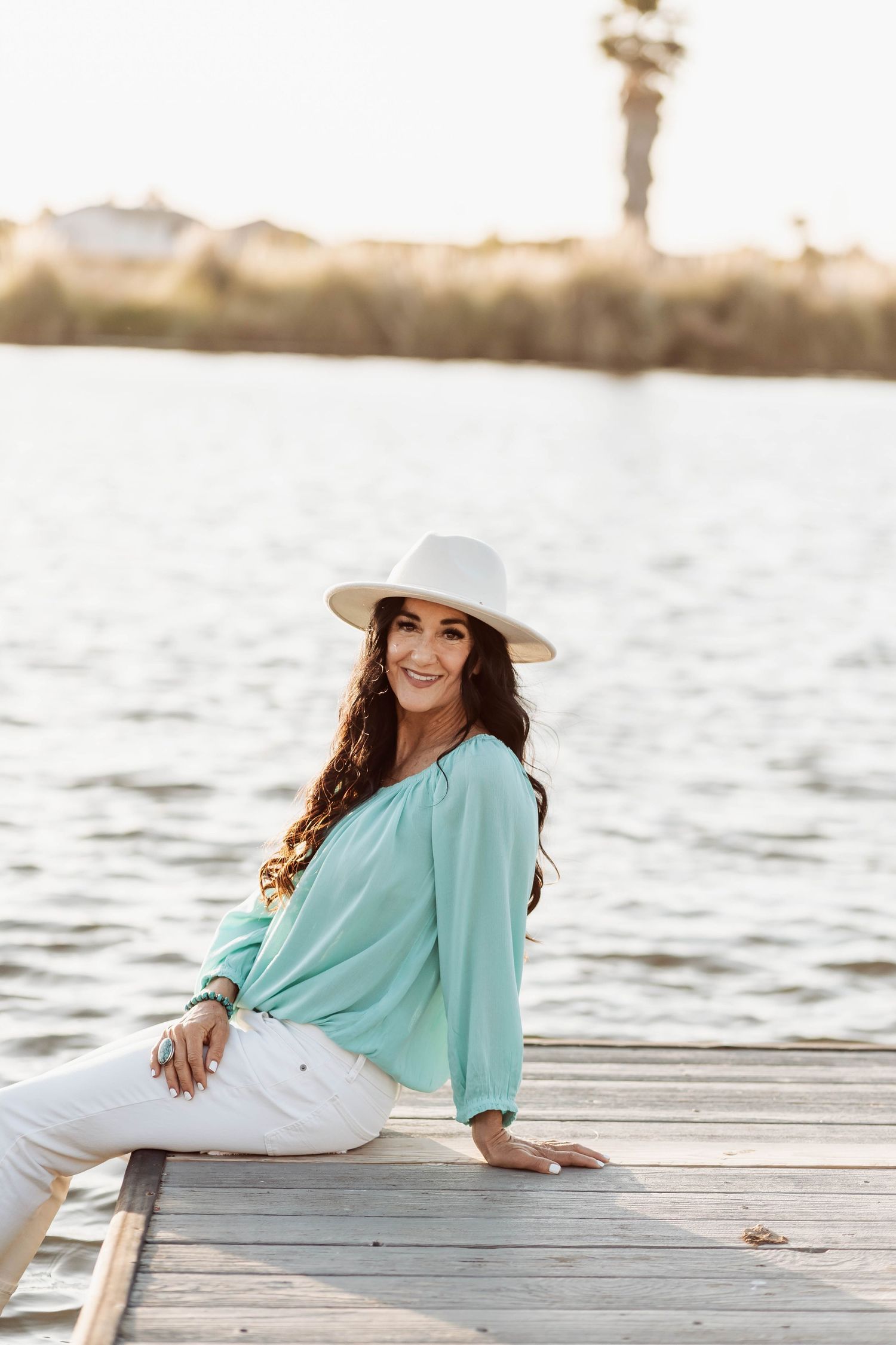 Woman in turquoise top and white pants sitting on a dock by the water at sunset.