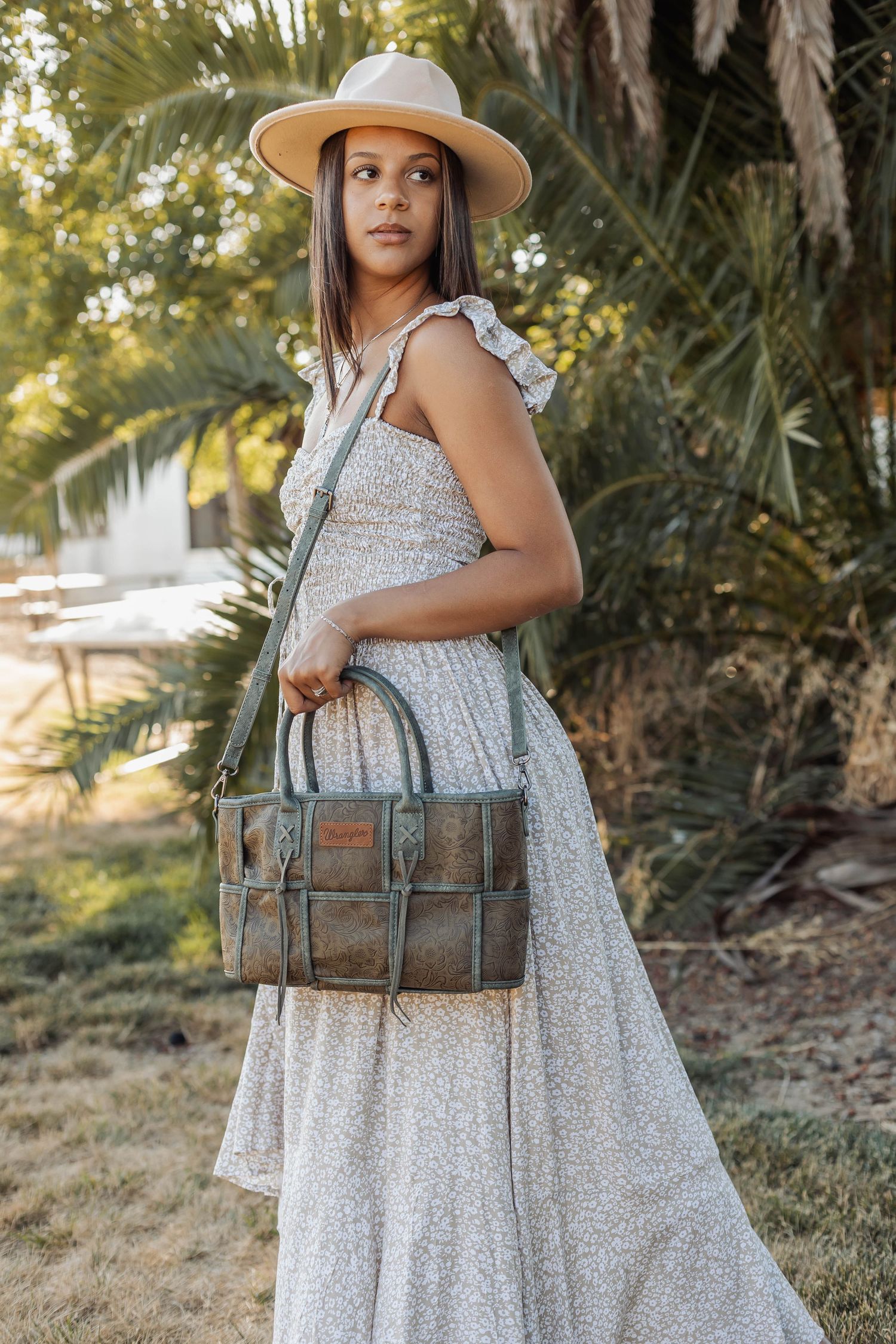 Vintage-style leather duffle bag paired with white dress and tan hat in garden setting.