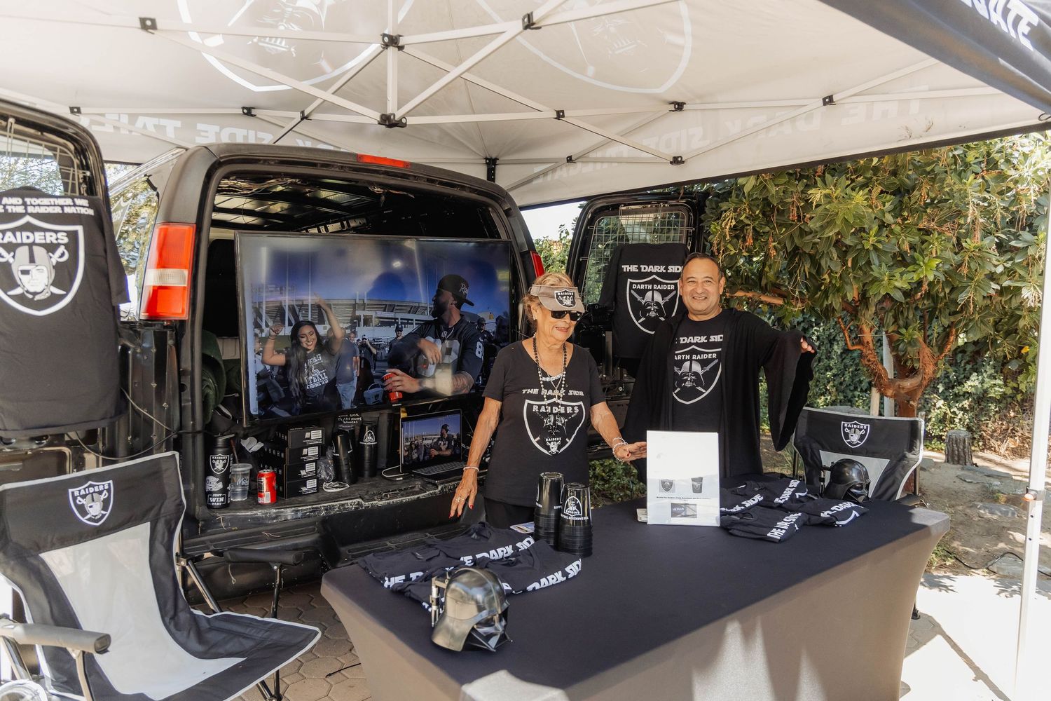 Two Raiders fans smile at a decorated tailgate booth with team merchandise and a van at an outdoor NFL event.
