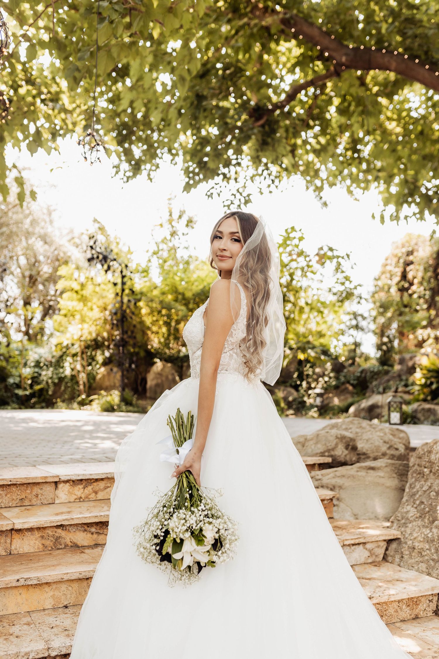 A bride in a flowing white ball gown poses on stone steps under green foliage with her wedding bouquet.
