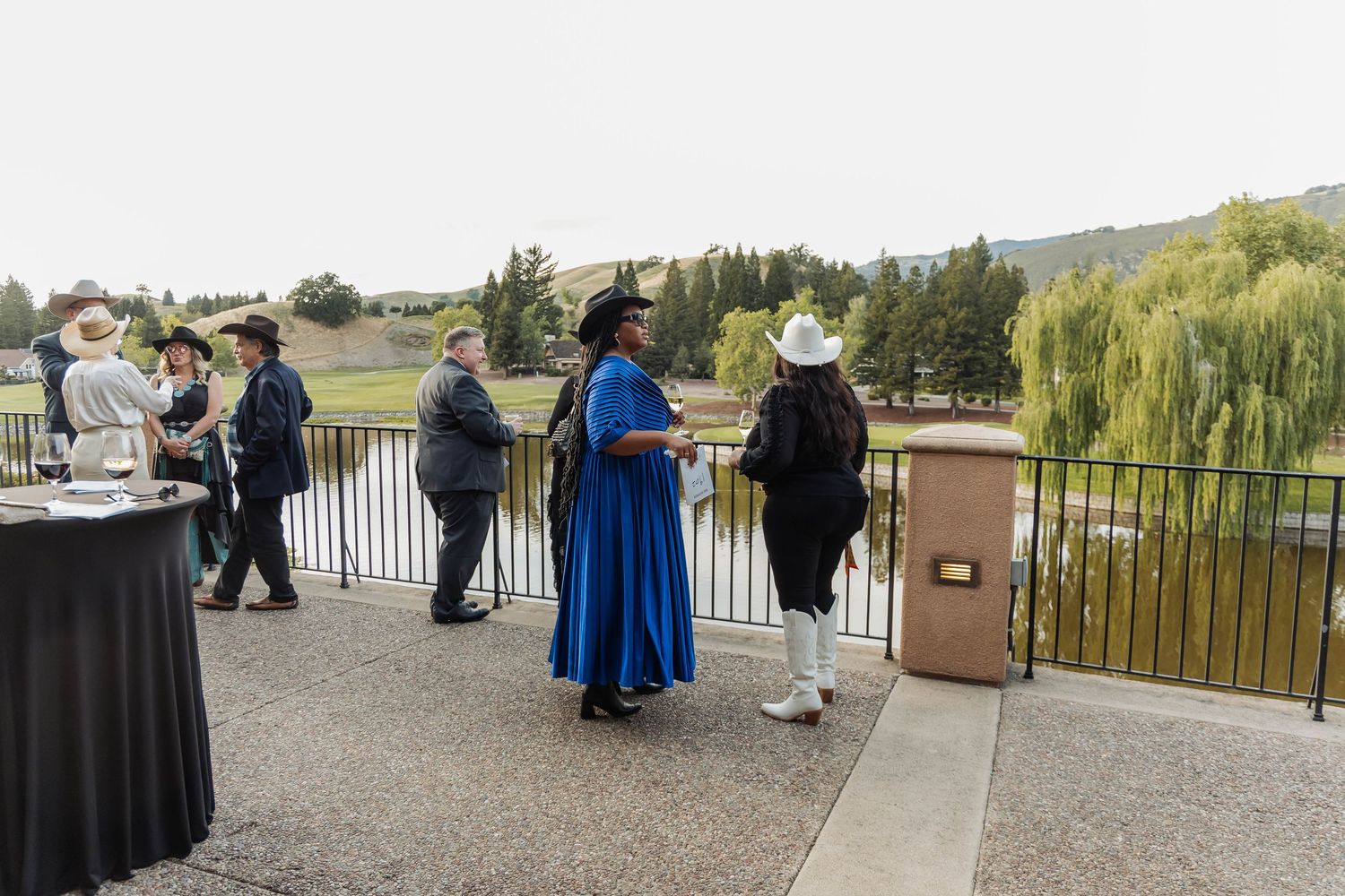 Attendees in formal Western attire socialize on an outdoor terrace overlooking a scenic green landscape.