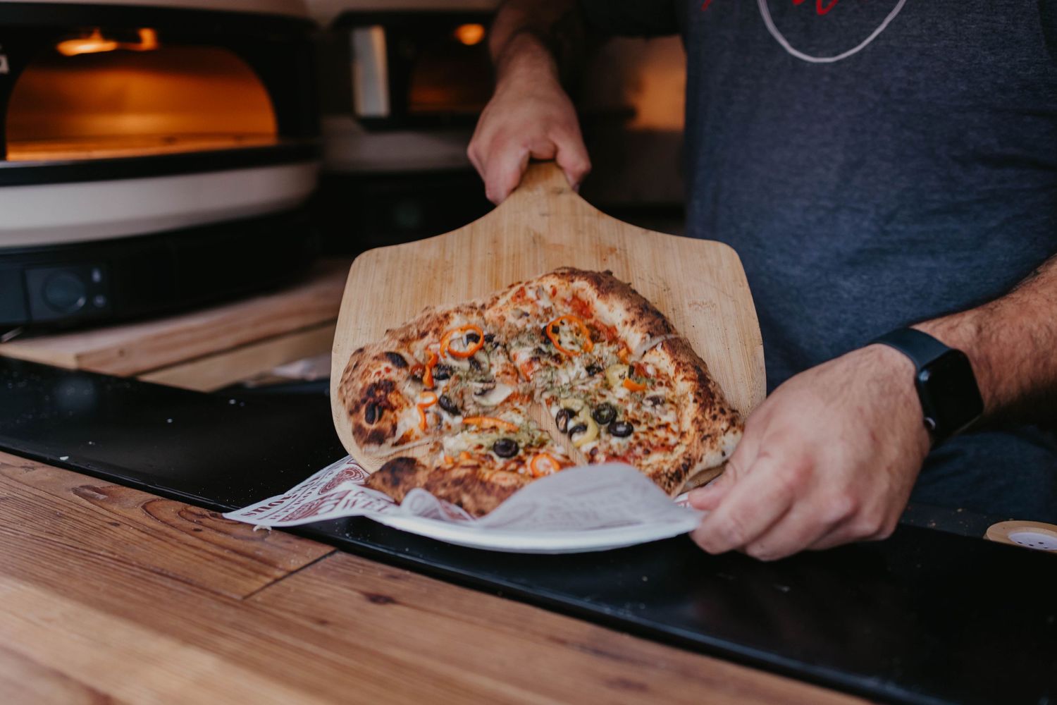 Pizza being served on a wooden table with dark kitchen decor in the background.