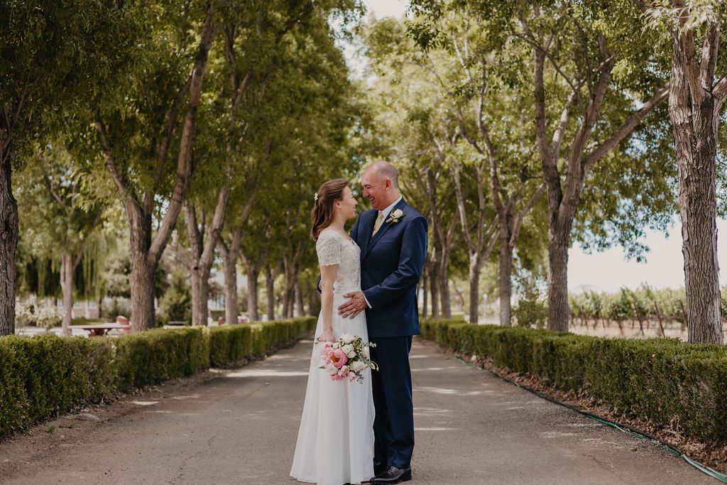 Wedding couple embraces on scenic vineyard path surrounded by mature trees and hedges.