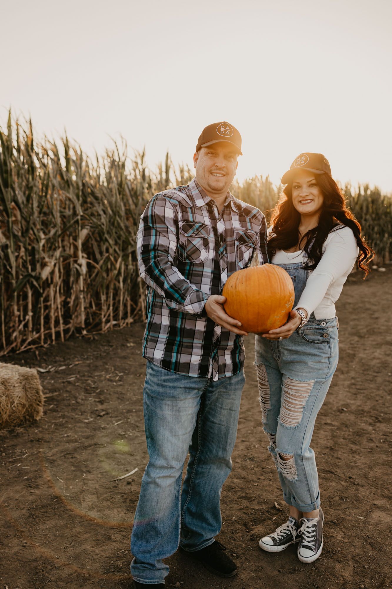 Couple holding pumpkin together in front of corn field during sunset.