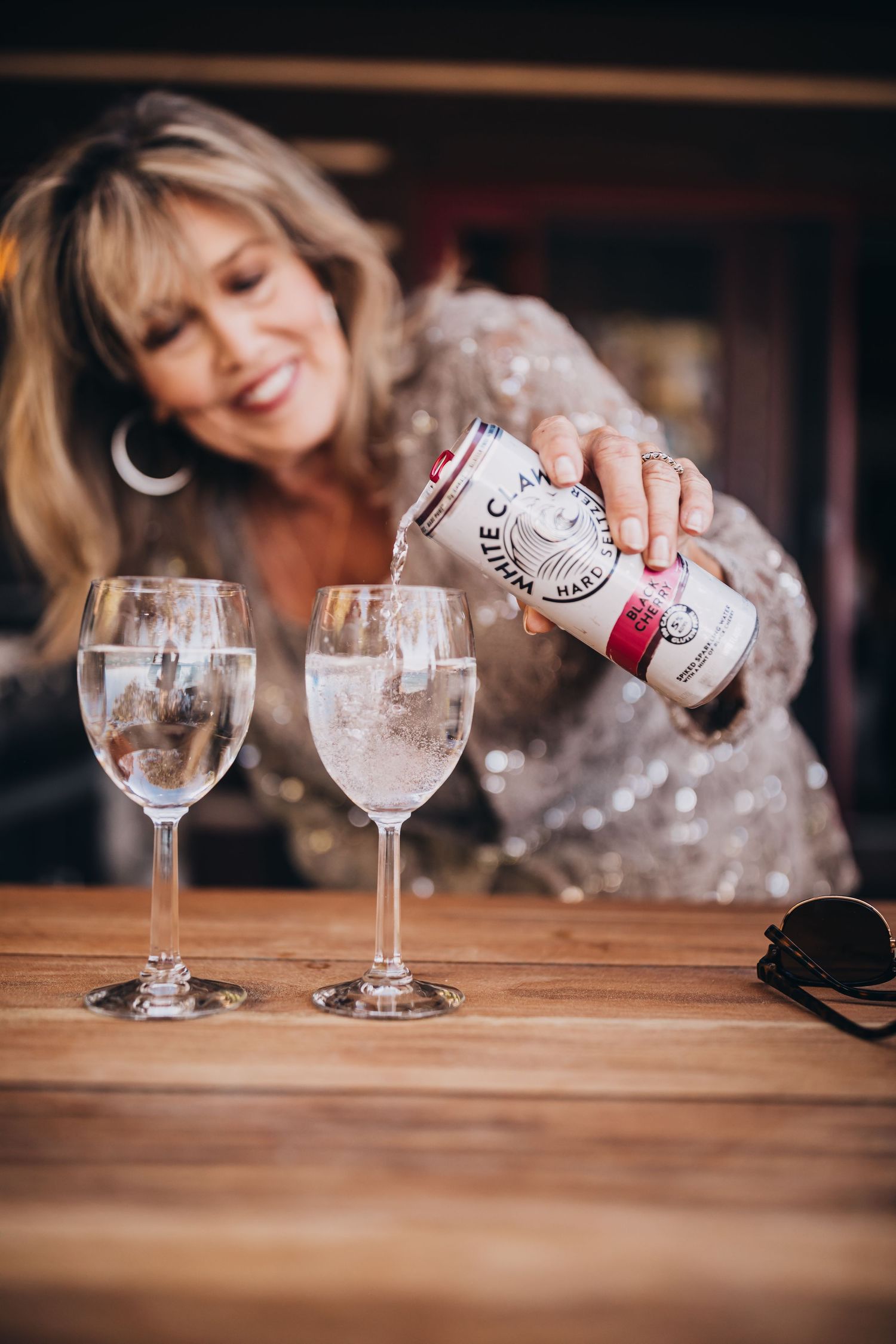 Someone pouring a canned beverage into wine glasses on a wooden table.