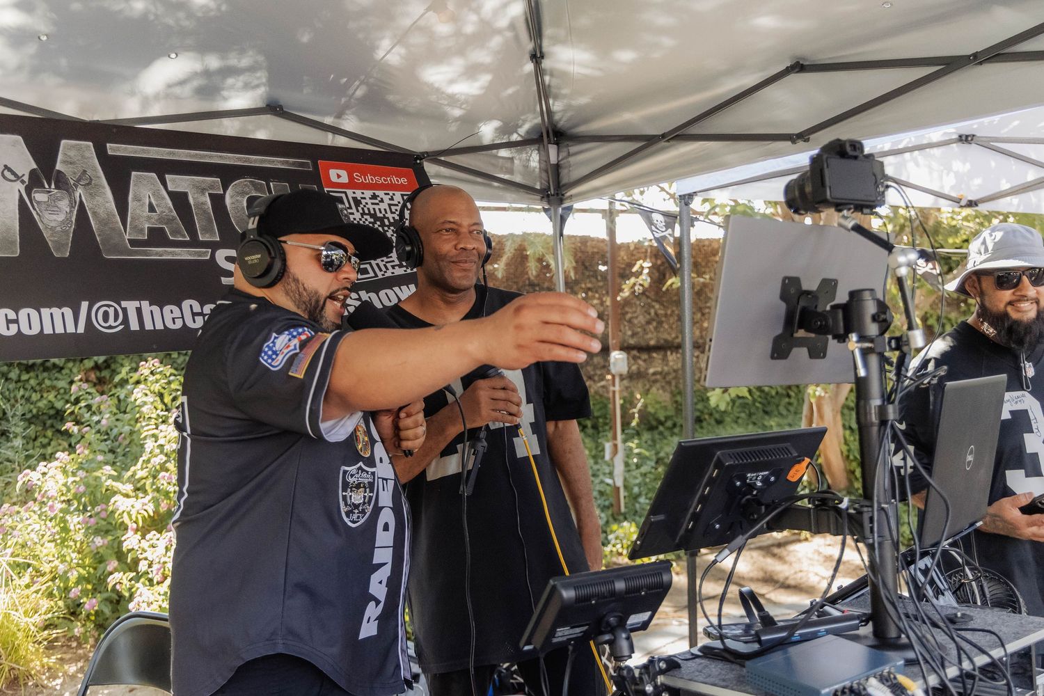 Two men operating DJ equipment and microphones under a tent at an outdoor Raiders fan event.