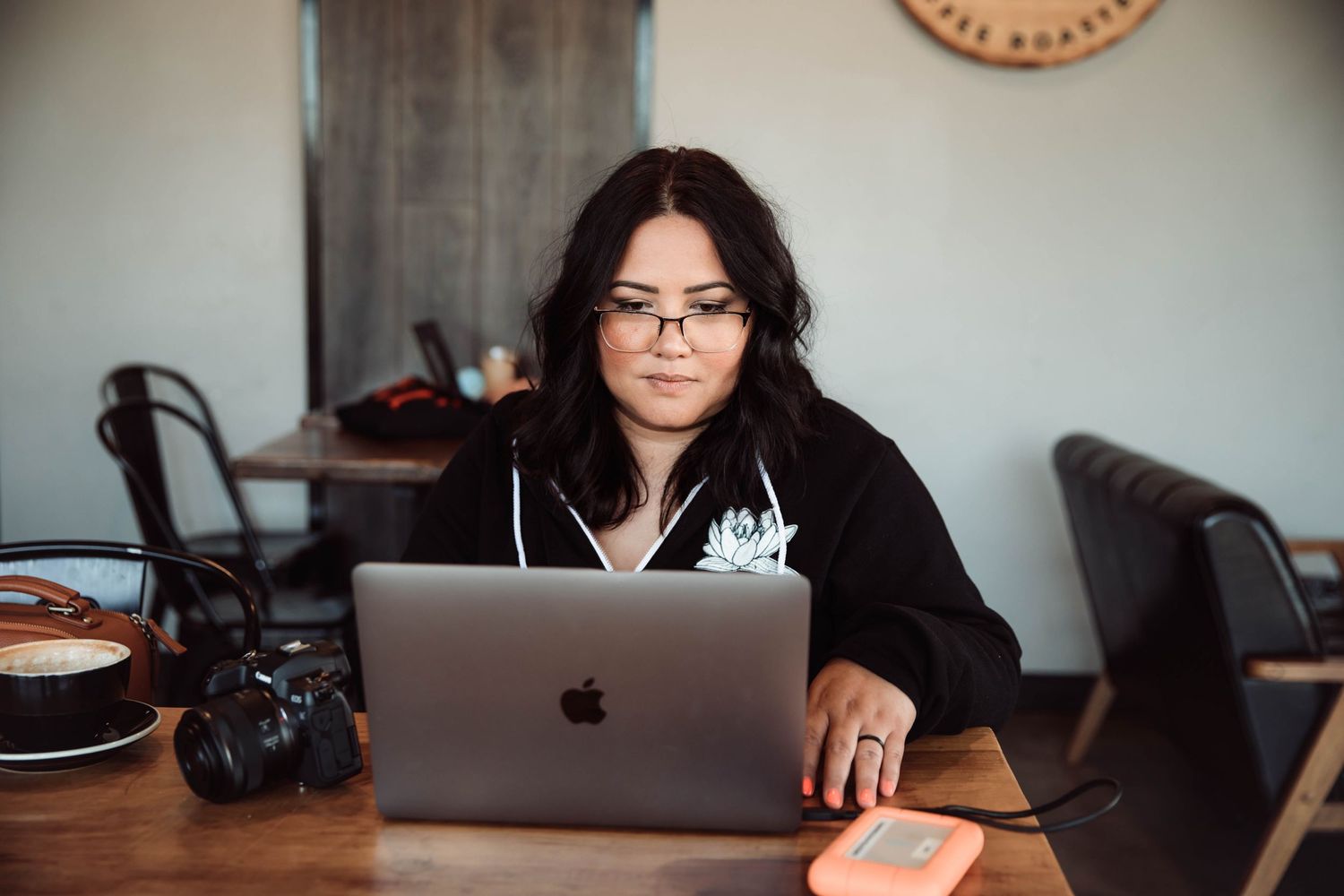 Woman with glasses working on MacBook laptop at wooden table in cafe with camera and coffee nearby.