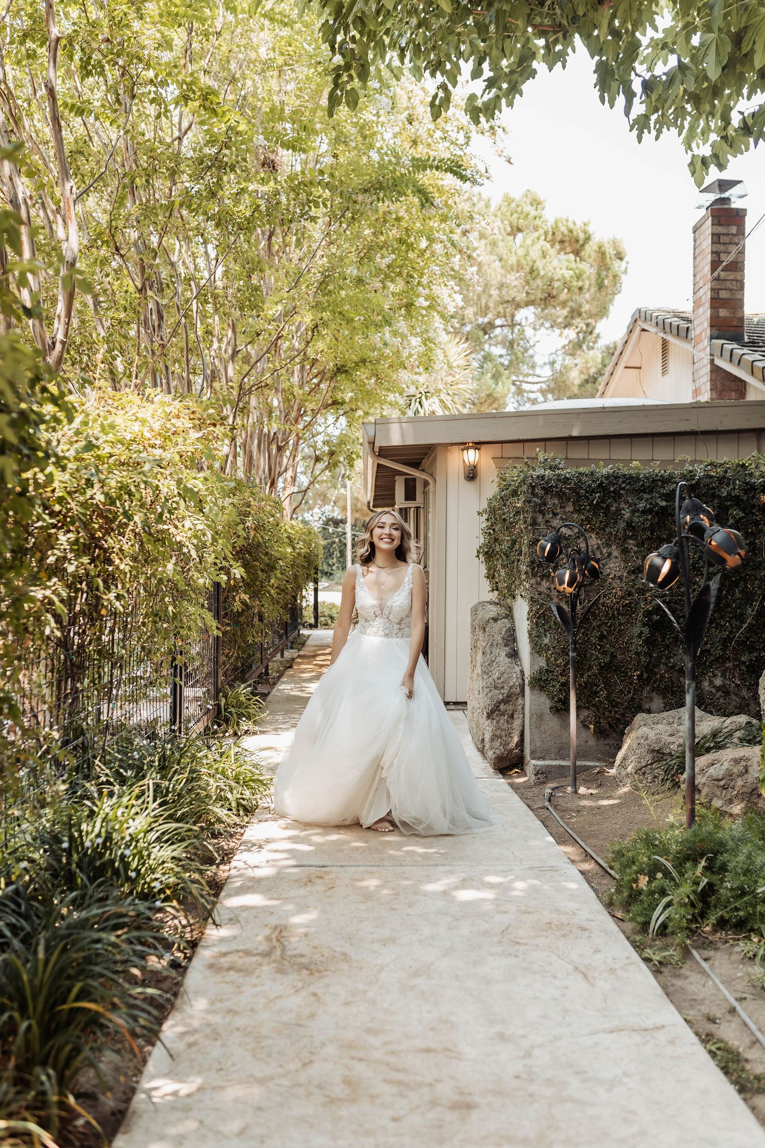 A bride in a princess ballgown walks along a sunlit garden pathway surrounded by lush greenery.