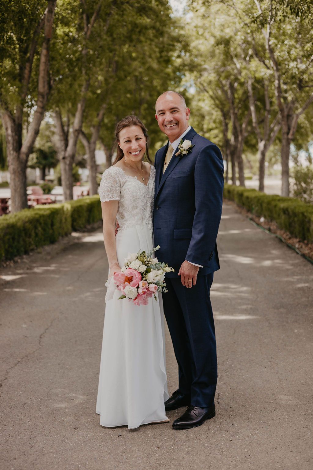 Newlyweds pose for portrait on tree-lined path with pink floral bouquet.