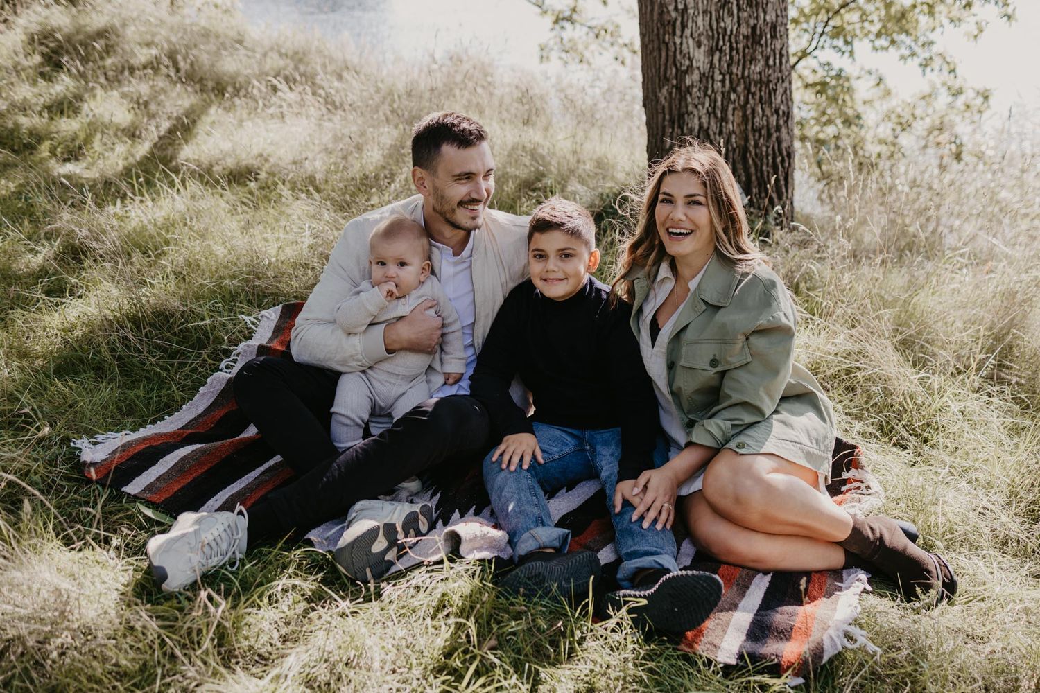 Casual family portrait taken outdoors with sunlit grass and trees in background.