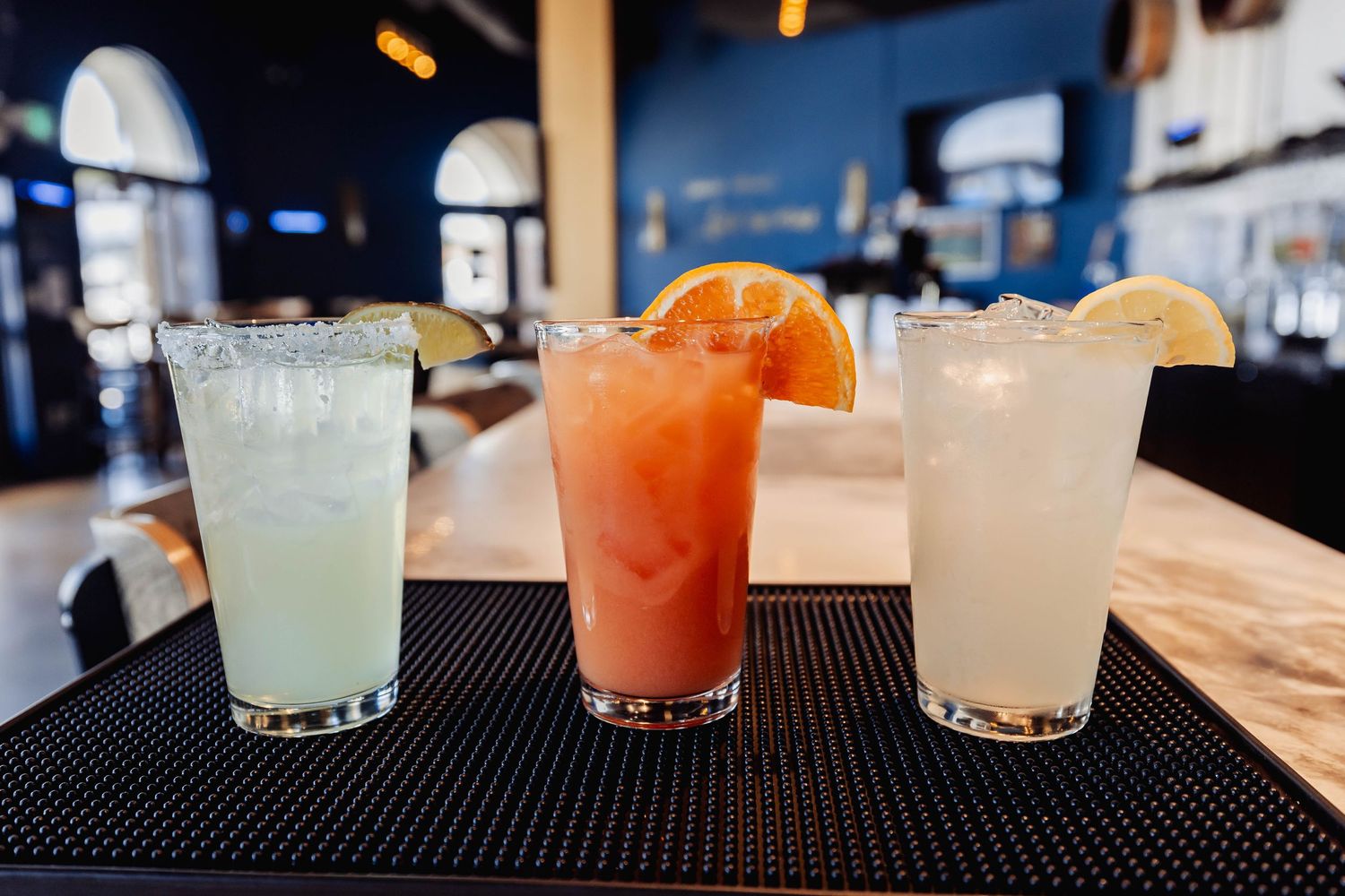 Row of colorful cocktails lined up on black bar mat in dimly lit setting.
