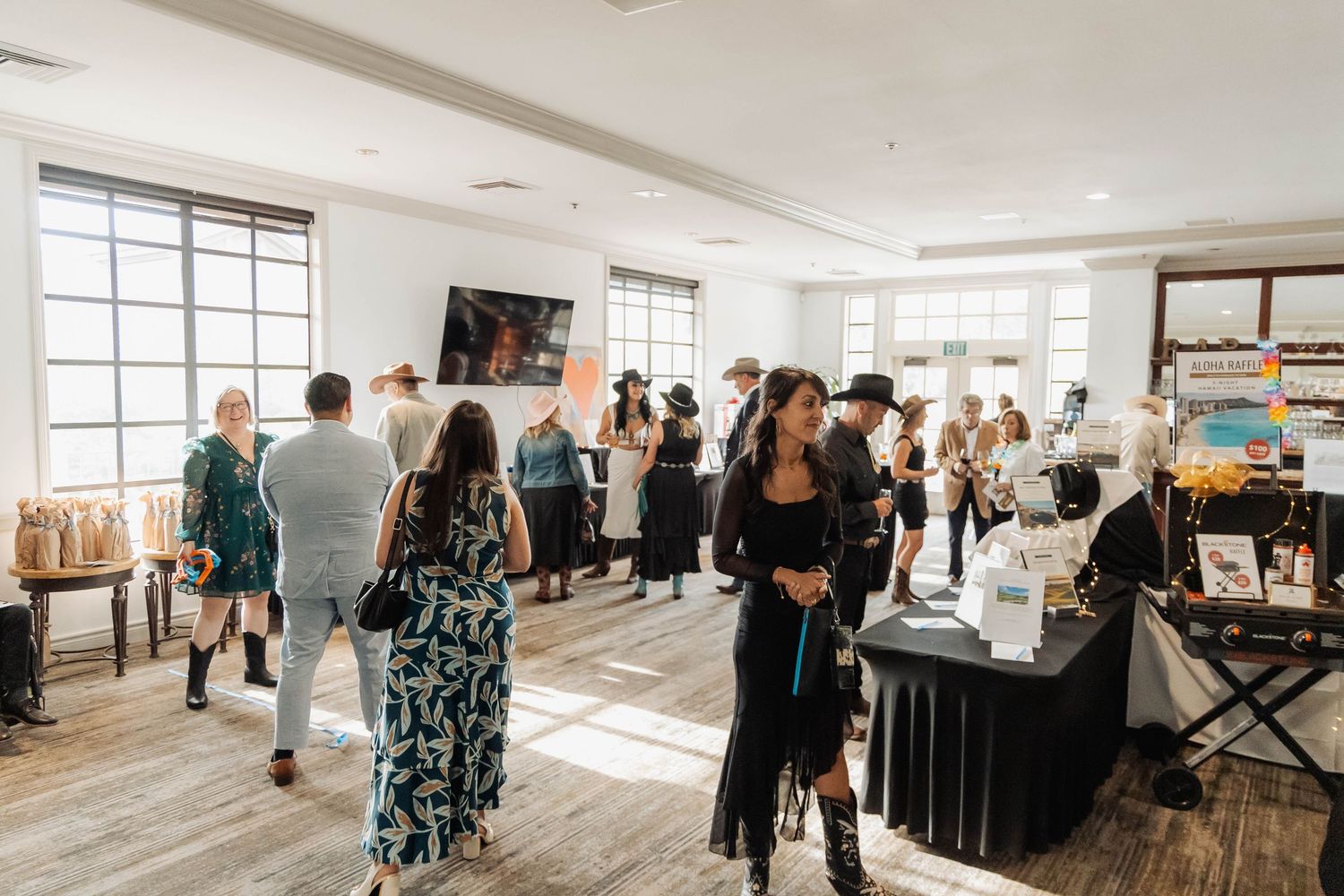 Guests browse vendor tables and socialize in a sunlit modern event space during a daytime gathering.