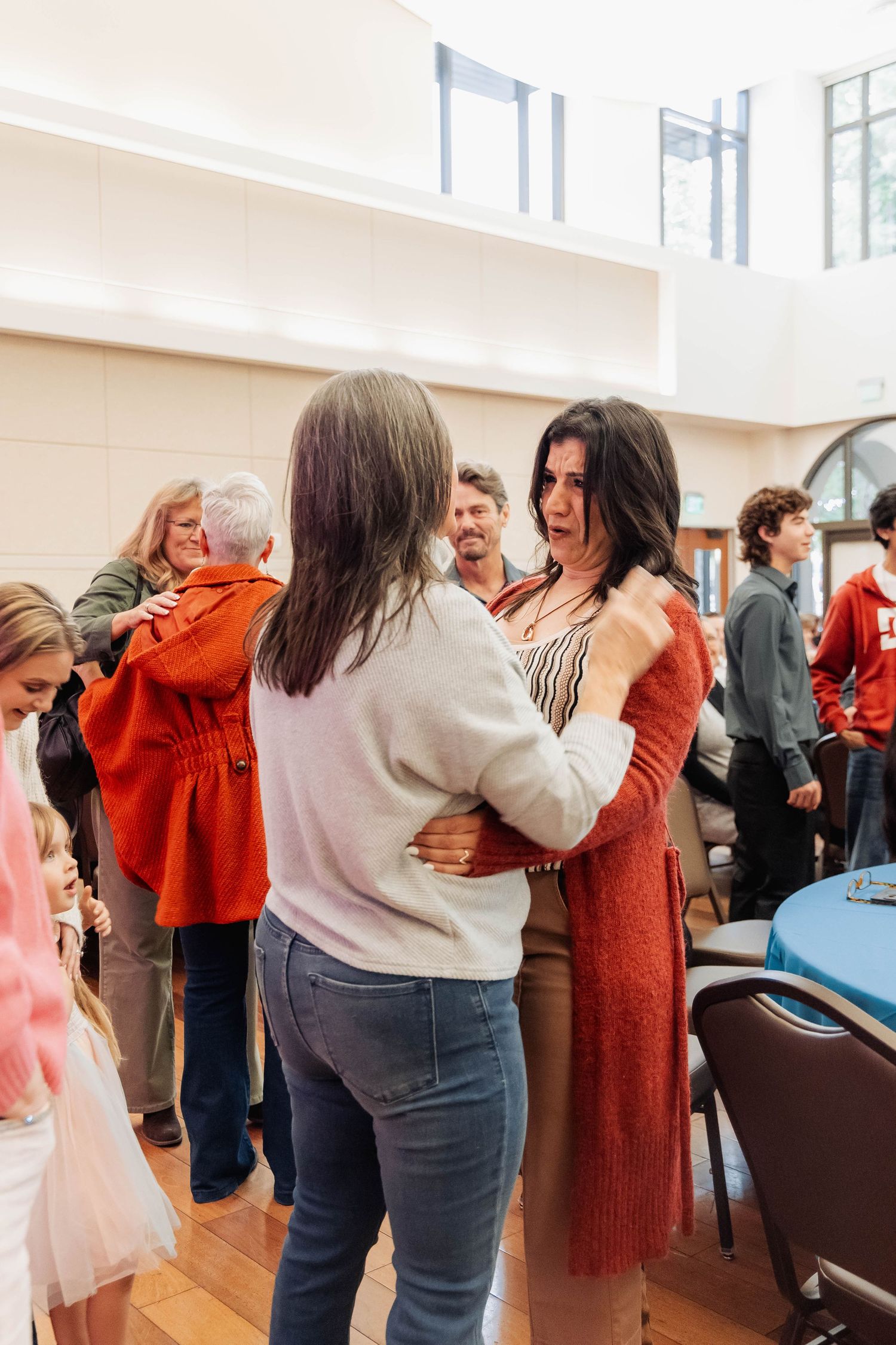 Two women embracing warmly in a bright community hall surrounded by other people socializing in the background.