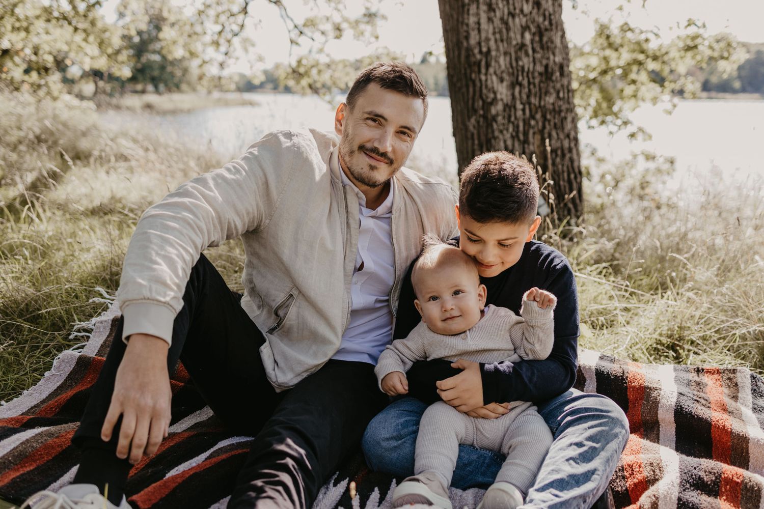 A family relaxing together on a picnic blanket under a tree in a sunlit field.