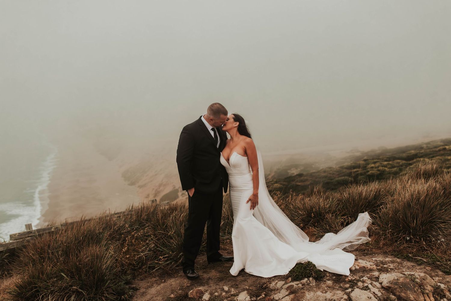 A newlywed couple embraces on a coastal cliff with dramatic fog in the background.