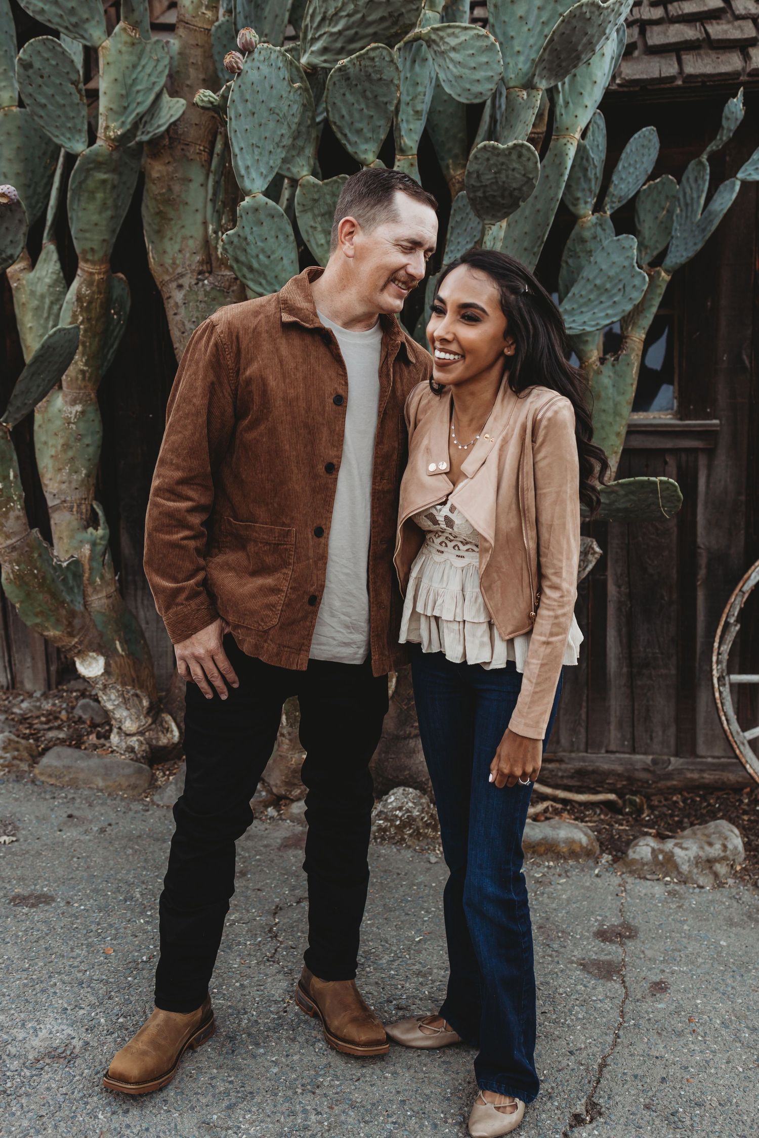 Couple sharing intimate moment in front of large desert cactus and rustic building.