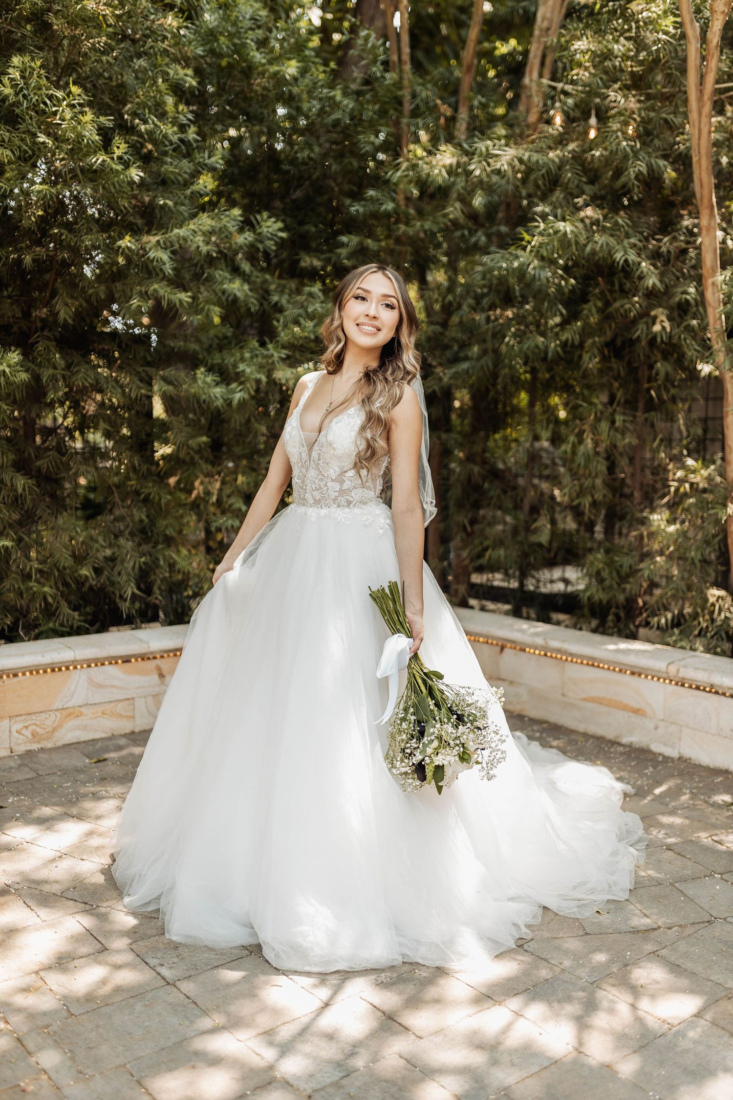 Sunlit garden bridal portrait featuring a flowing white gown with lace bodice and floral bouquet.