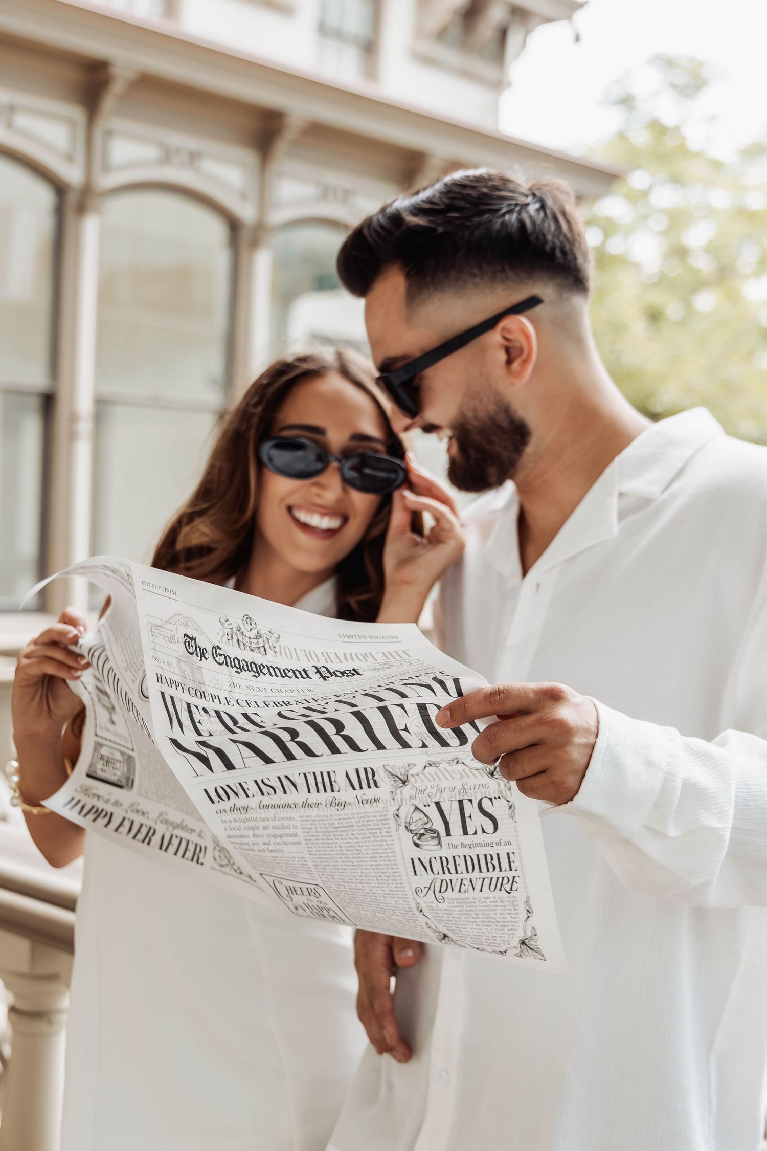 Stylish couple in white outfits smiling while holding wedding announcement newspaper on balcony.
