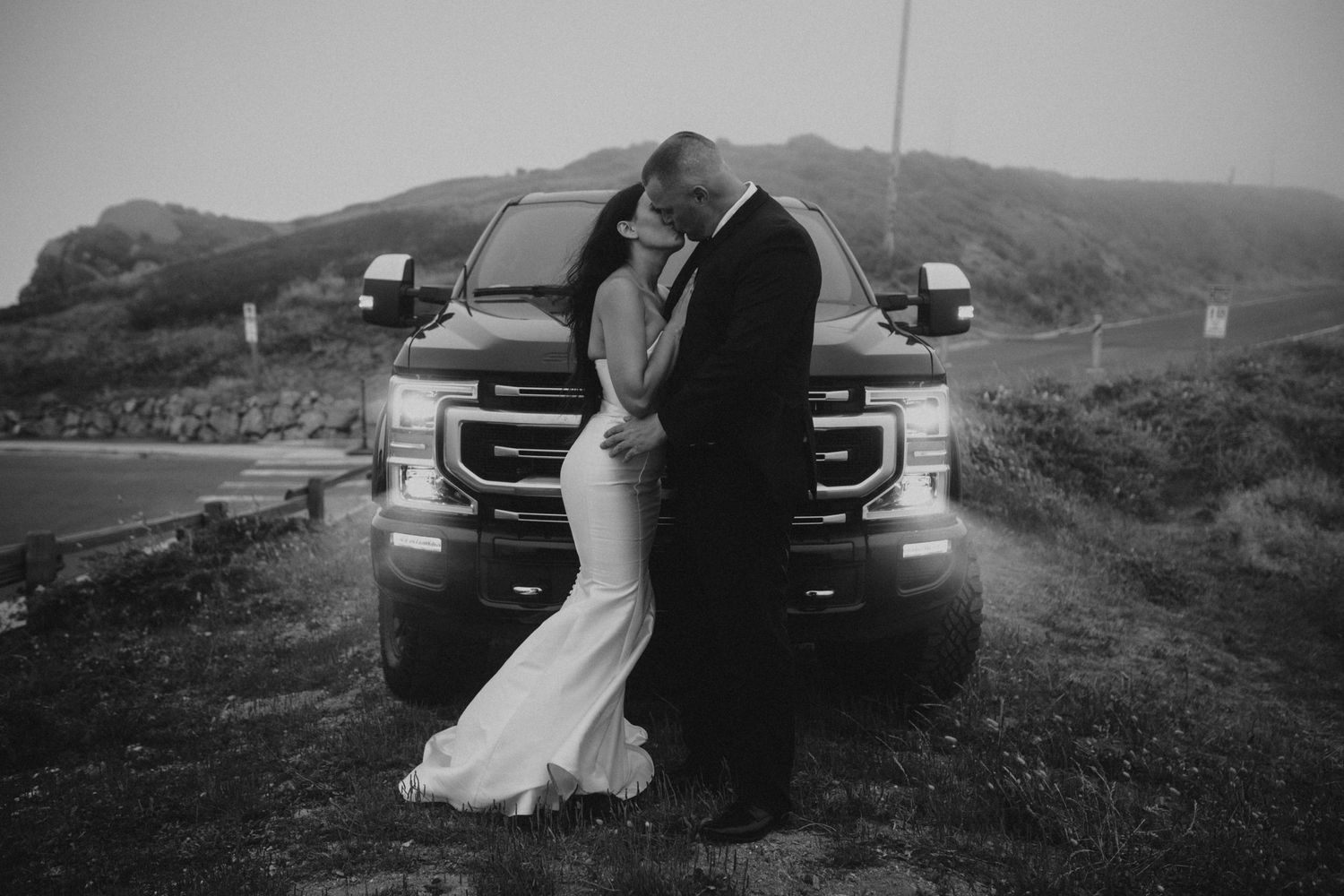 A black and white wedding portrait with a couple standing against a truck on a misty hill.