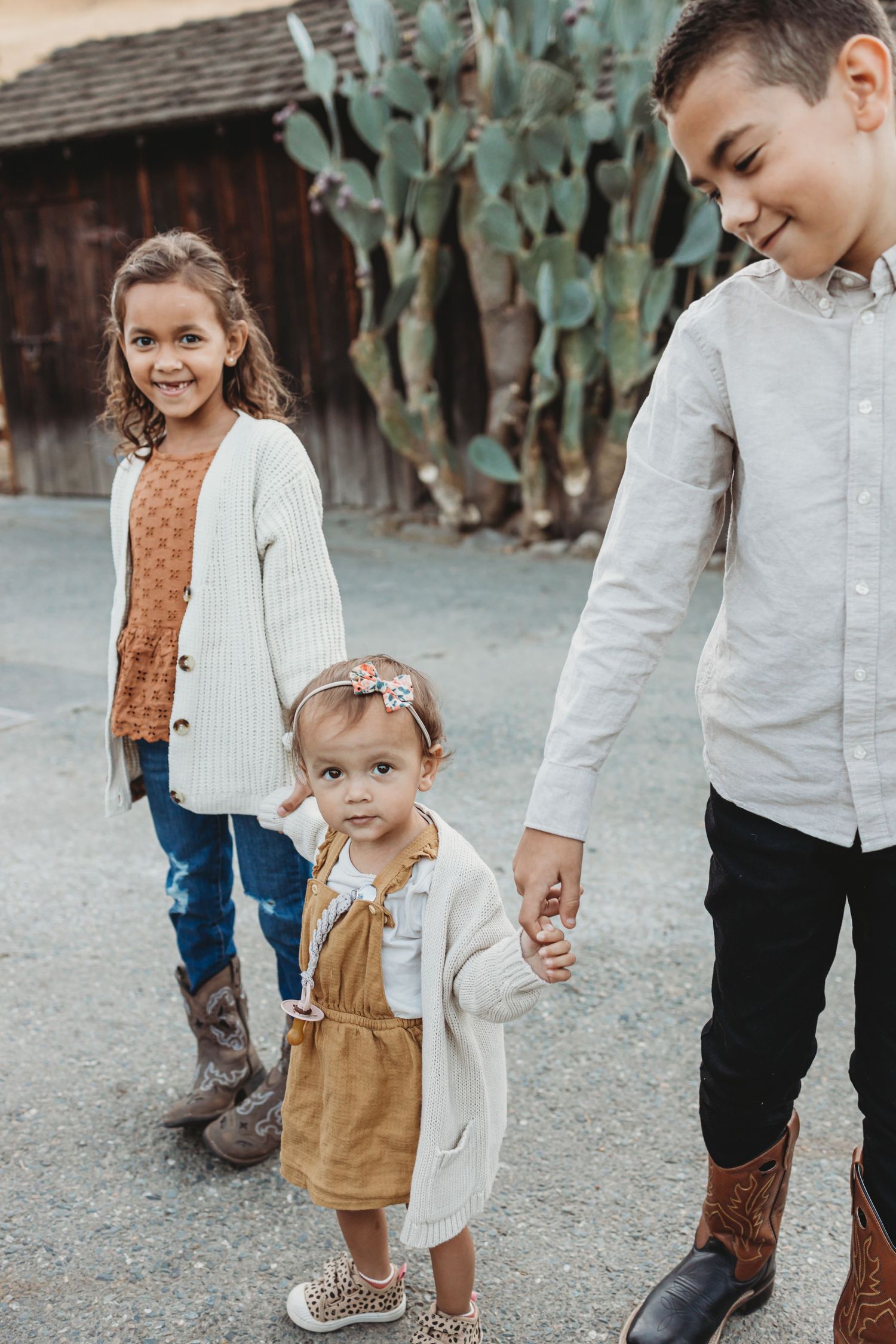 Three children standing together on path with large cactus and rustic building behind them.