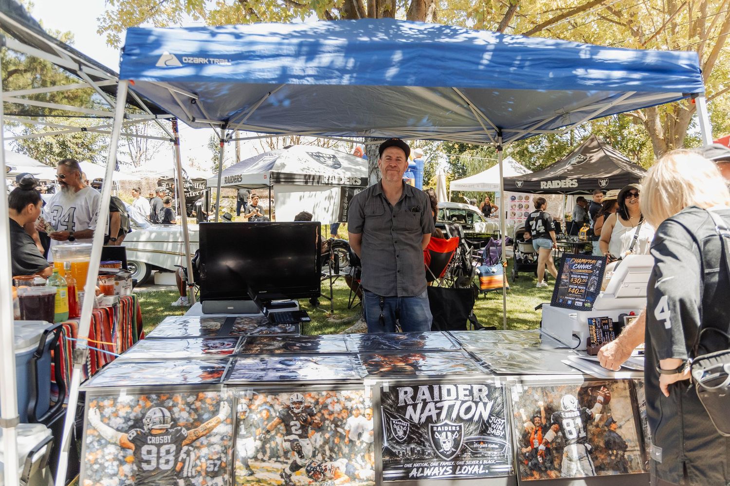 Vendor at outdoor market standing behind a Raider Nation merchandise booth with sports memorabilia under a blue canopy.