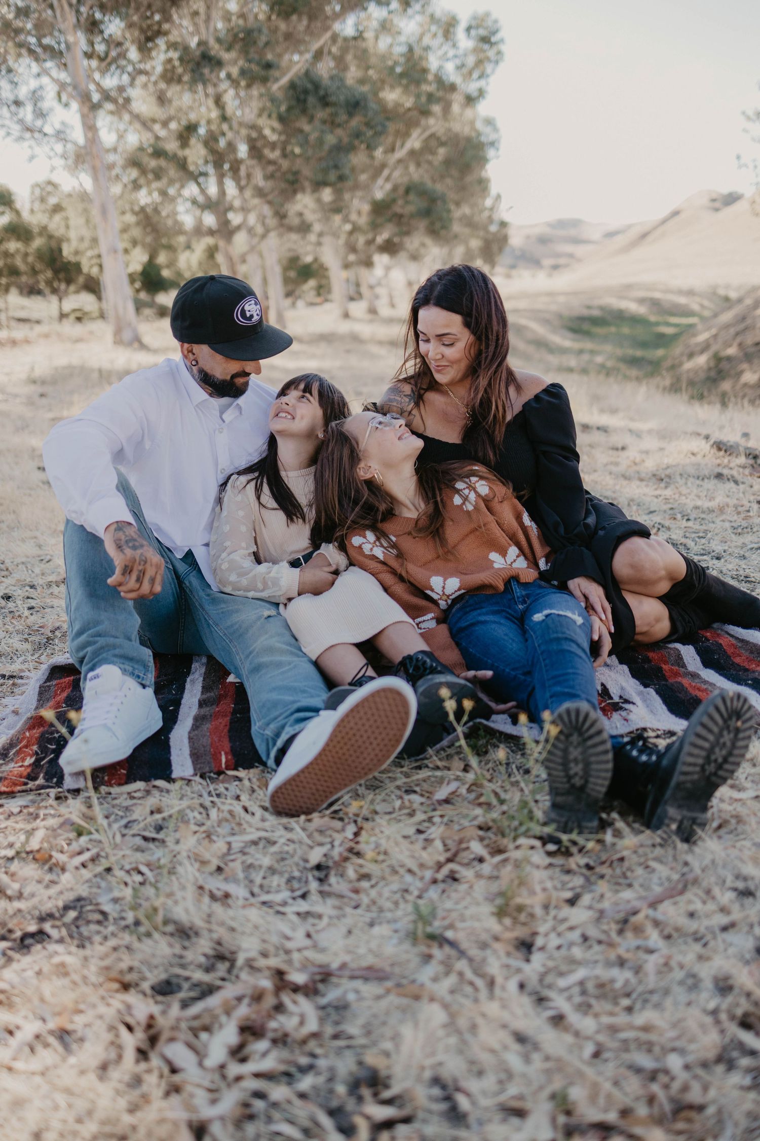 Family of four laughing together on a blanket outdoors in a golden field with trees and hills.
