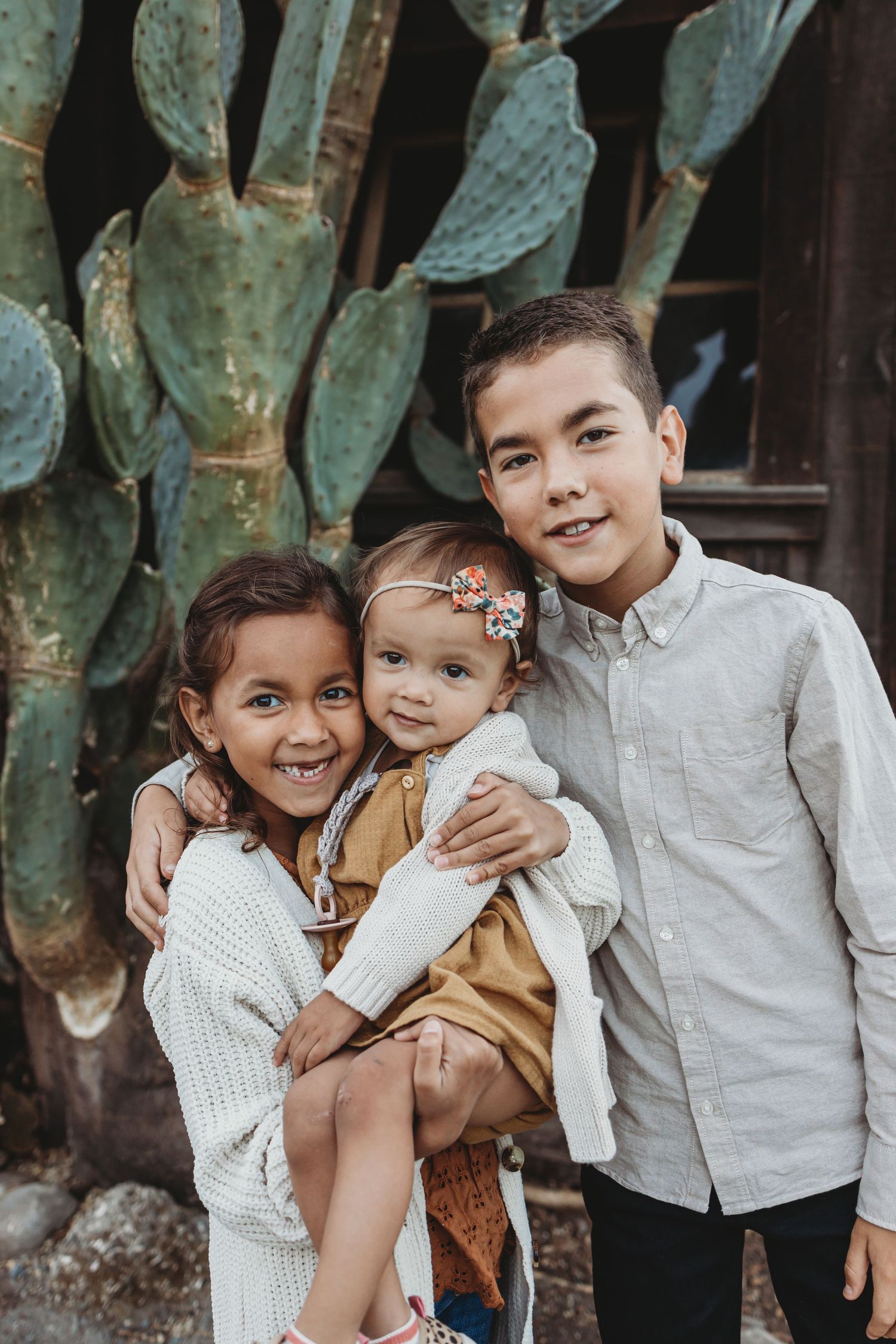 Three siblings pose together in front of large cactus plant and rustic building.