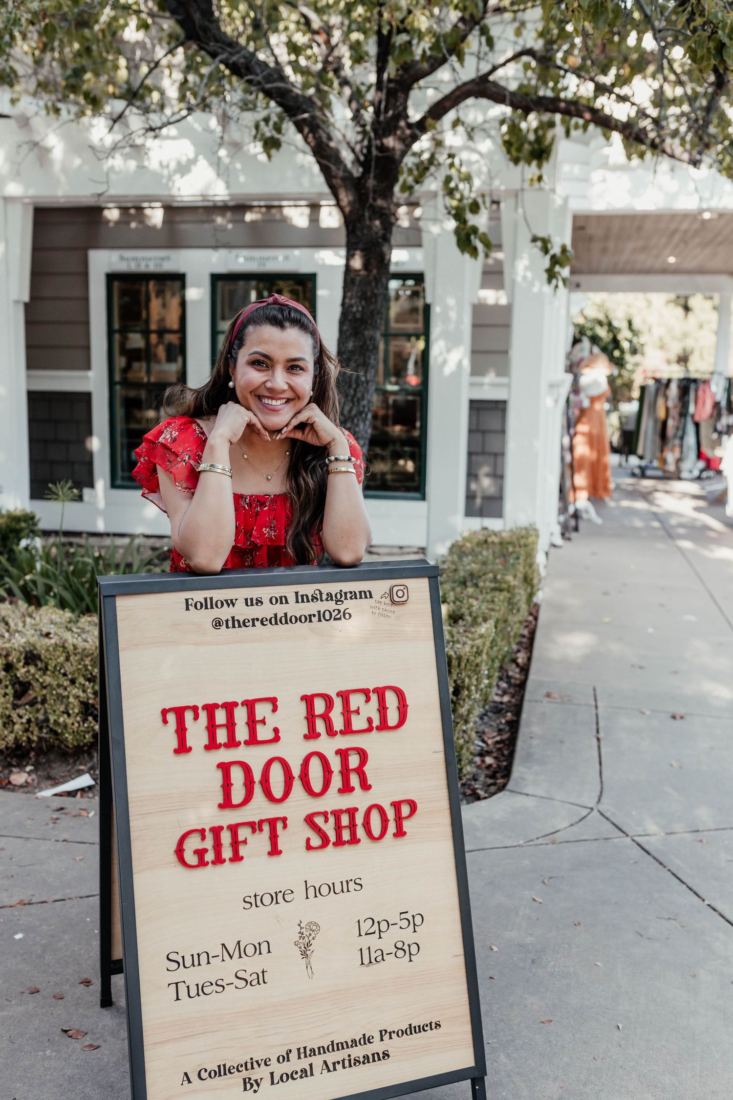 A store hours sign for The Red Door Gift Shop displayed outdoors under a shady tree.
