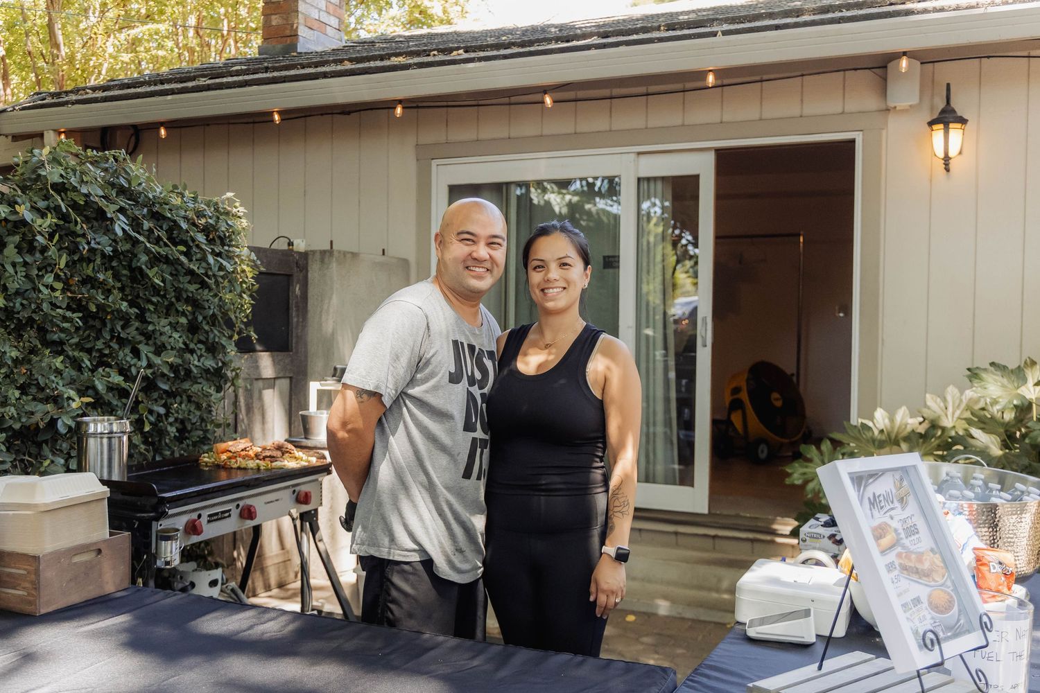 Couple smiling together outdoors near a grill at a backyard gathering beside a wooden home.