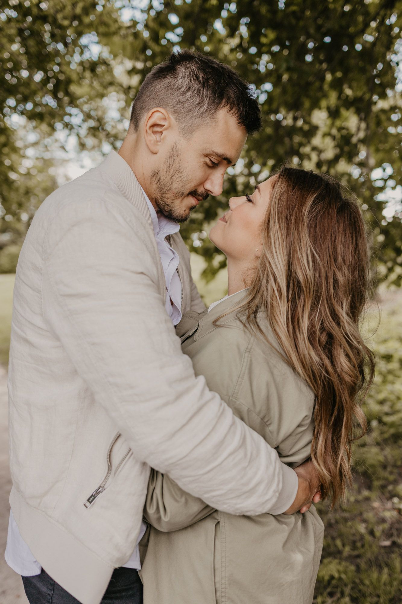 A romantic couple embraces outdoors on a sunny day surrounded by trees.